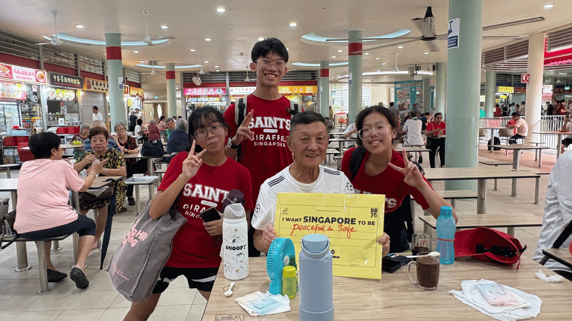 Inside an open-air hawker center, an elderly man in a white t-shirt sits at a table holding a yellow paper sign with handwritten text that reads "I WANT SINGAPORE TO BE peaceful & safe IN 2040." Three students wearing red "SAINTS GIRAFFES SINGAPORE" t-shirts surround him, posing cheerfully with peace signs. Water bottles, a small blue portable fan, and a glass of iced drink sit on the table in front of them, with hawker stalls and other patrons visible in the background.