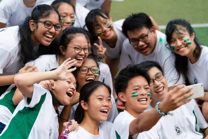 A group of smiling teens with green face paint in white shirts take a selfie together outdoors.
