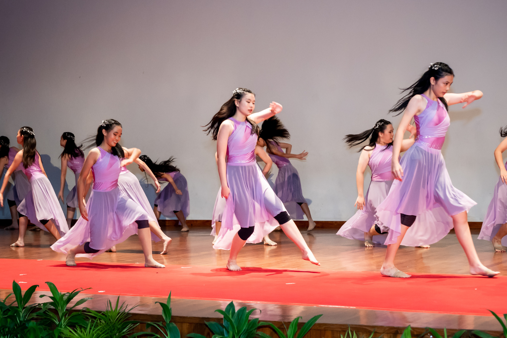 A group of young female dancers perform on stage, wearing matching lavender/purple flowing dresses with pink accents and black leggings underneath. Their hair is loose and flowing with floral hair accessories. They appear to be mid-movement in a synchronised contemporary or lyrical dance routine, barefoot on a wooden stage with a red carpet at the front. Potted plants are visible at the base of the stage.