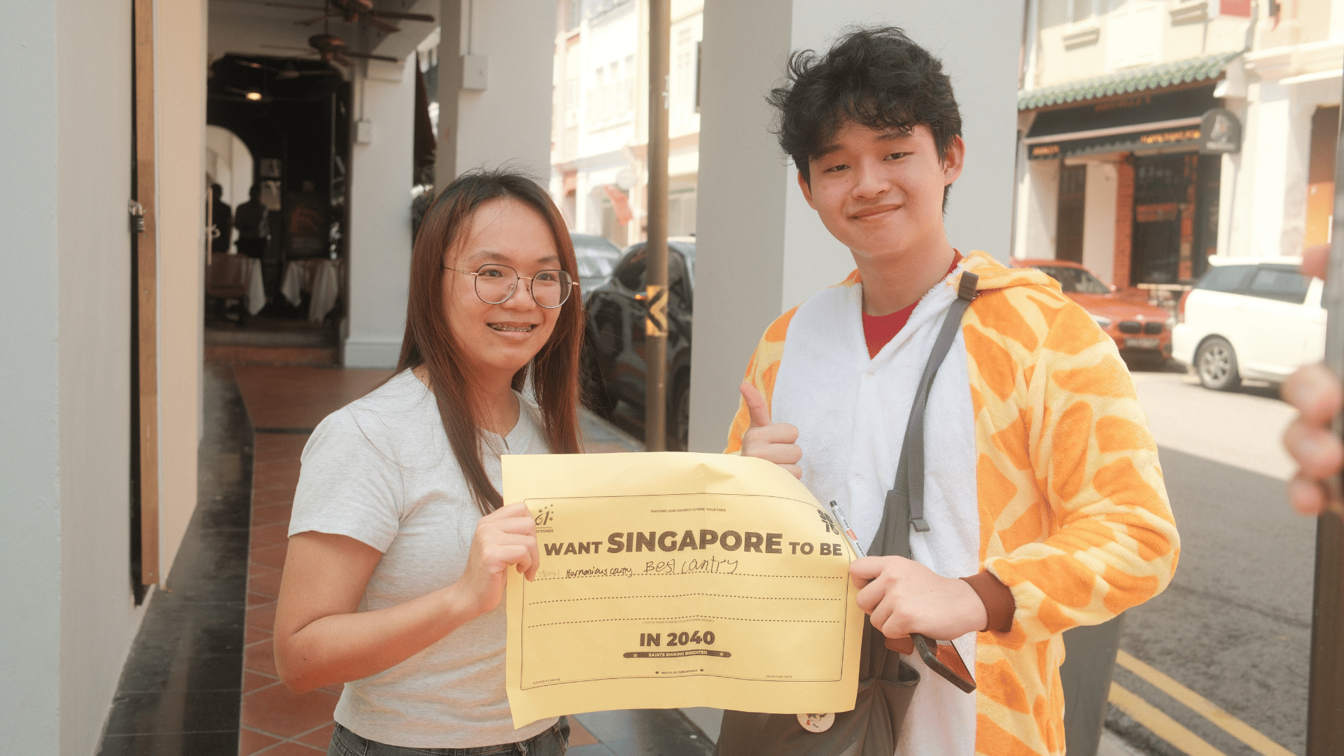 A woman wearing glasses and a light grey t-shirt stands beside a young man dressed in a hooded giraffe onesie along a covered walkway. Together, they hold up a yellow paper sign that reads "I WANT SINGAPORE TO BE..." with handwritten text filled in, above "IN 2040." The man gives a thumbs-up gesture while holding a marker, with a street lined with shophouses and parked cars visible in the background.