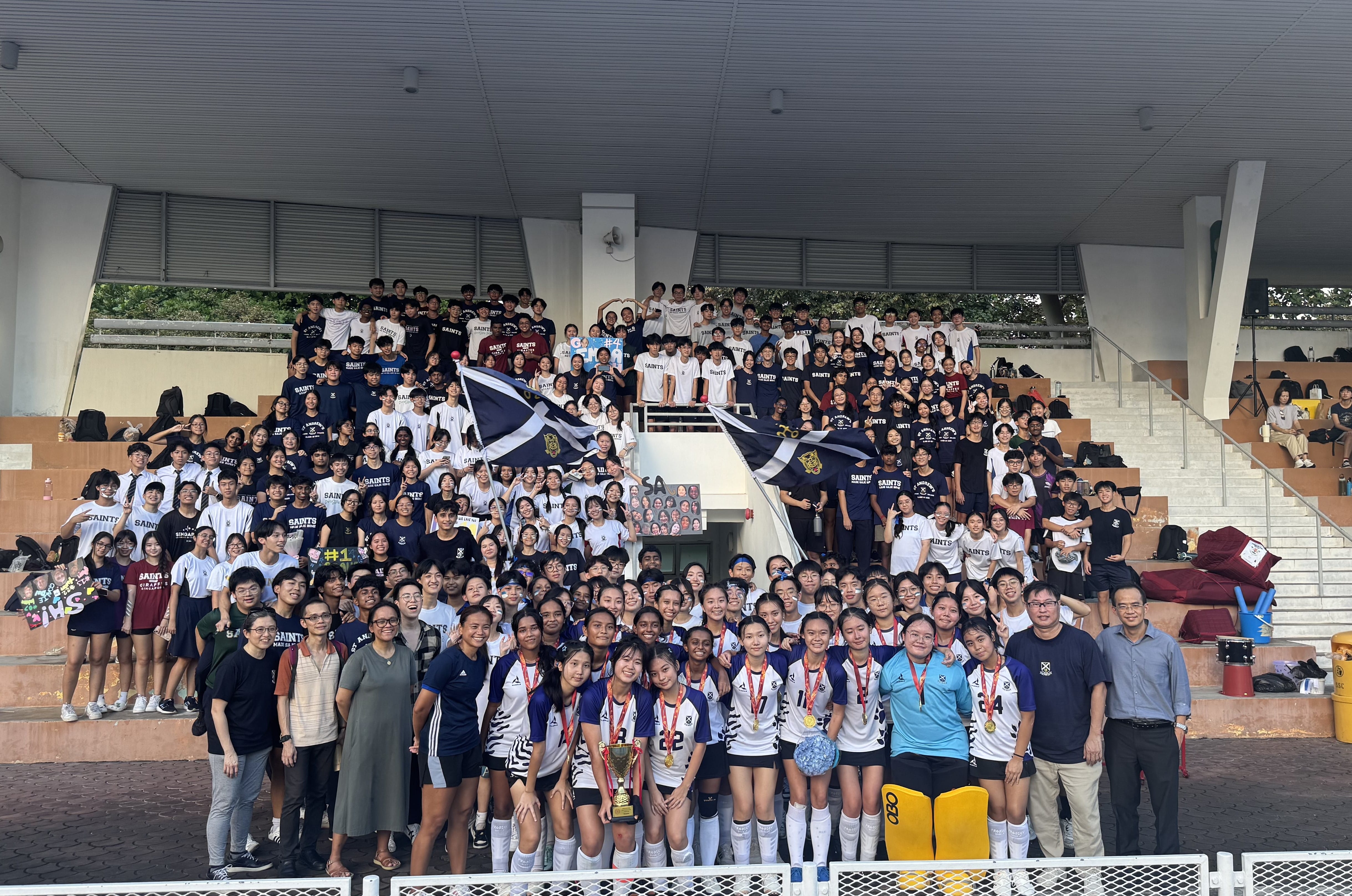 Large group of students in navy and white Saints uniforms posing on stadium bleachers, with athletes holding a trophy and medals.