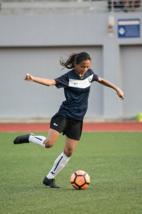 A young woman in a navy jersey and black shorts kicks an orange soccer ball on a green grass field.