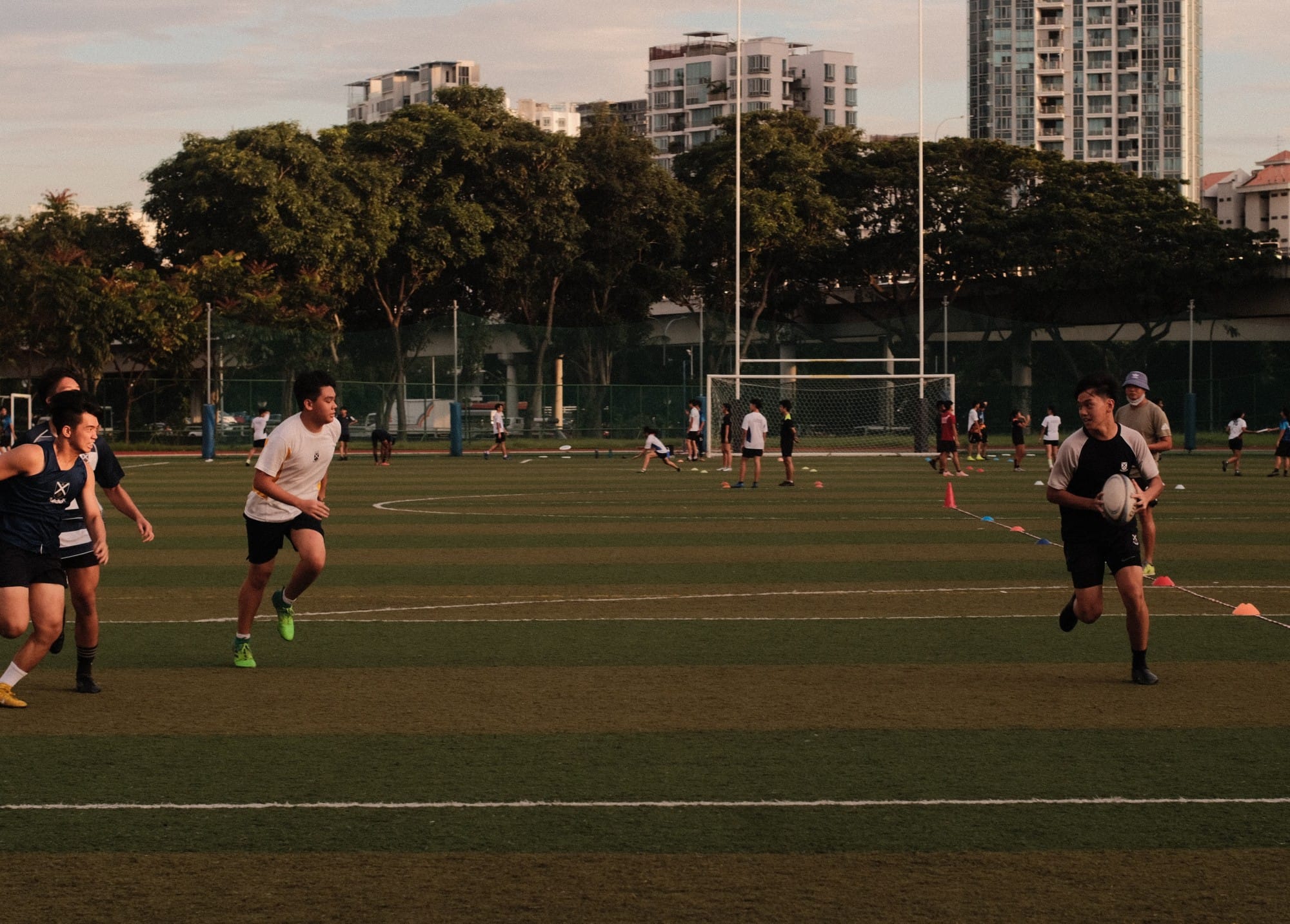 Students play touch rugby on a green field at dusk, with one player carrying the ball while others give chase, set against a backdrop of trees and residential high-rise buildings.