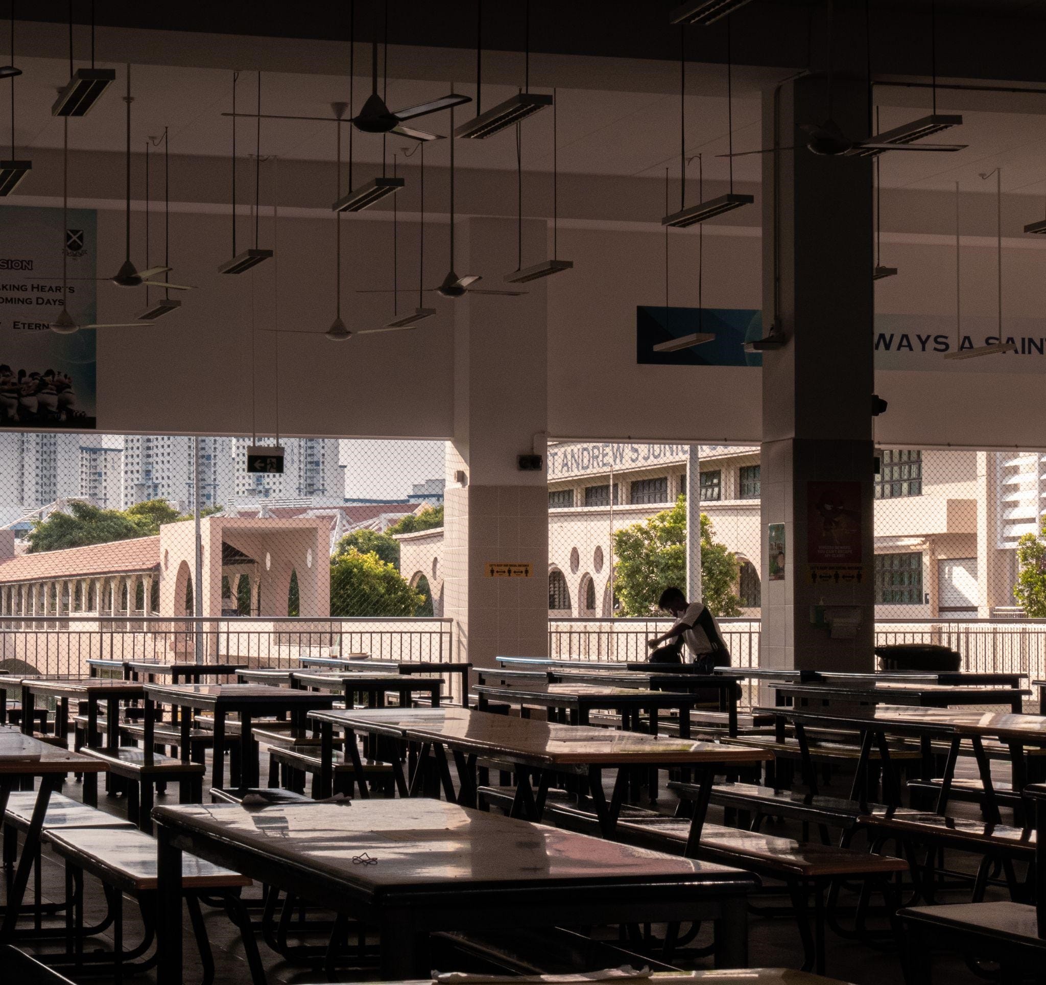 A lone student sits at a canteen table in a quiet, empty school canteen, with the school building visible in the background. A sign on the wall reads 'Always a Saint'.