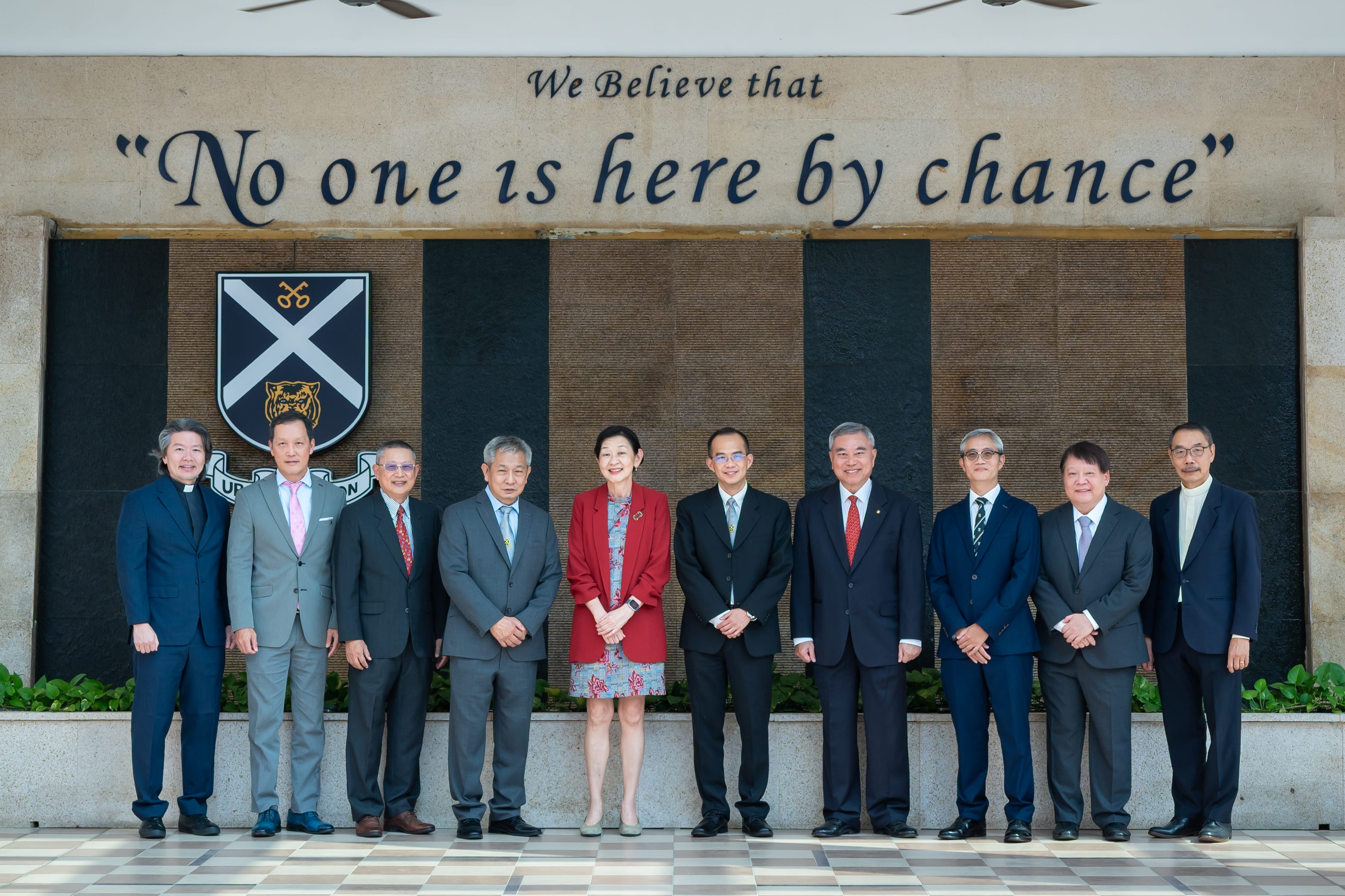 Nine formally dressed adults pose under wall text, No one is here by chance, beside a shield crest.