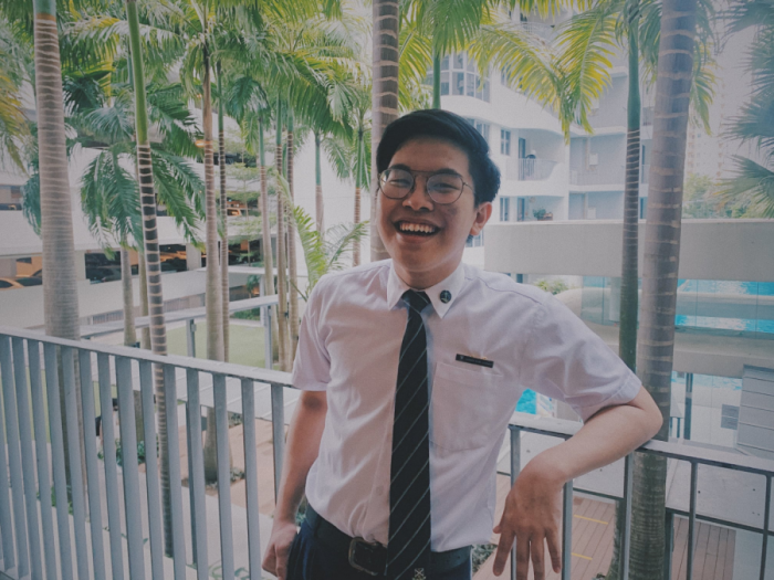 Young man in white shirt and dark tie laughing on a balcony with palm trees and buildings behind him.