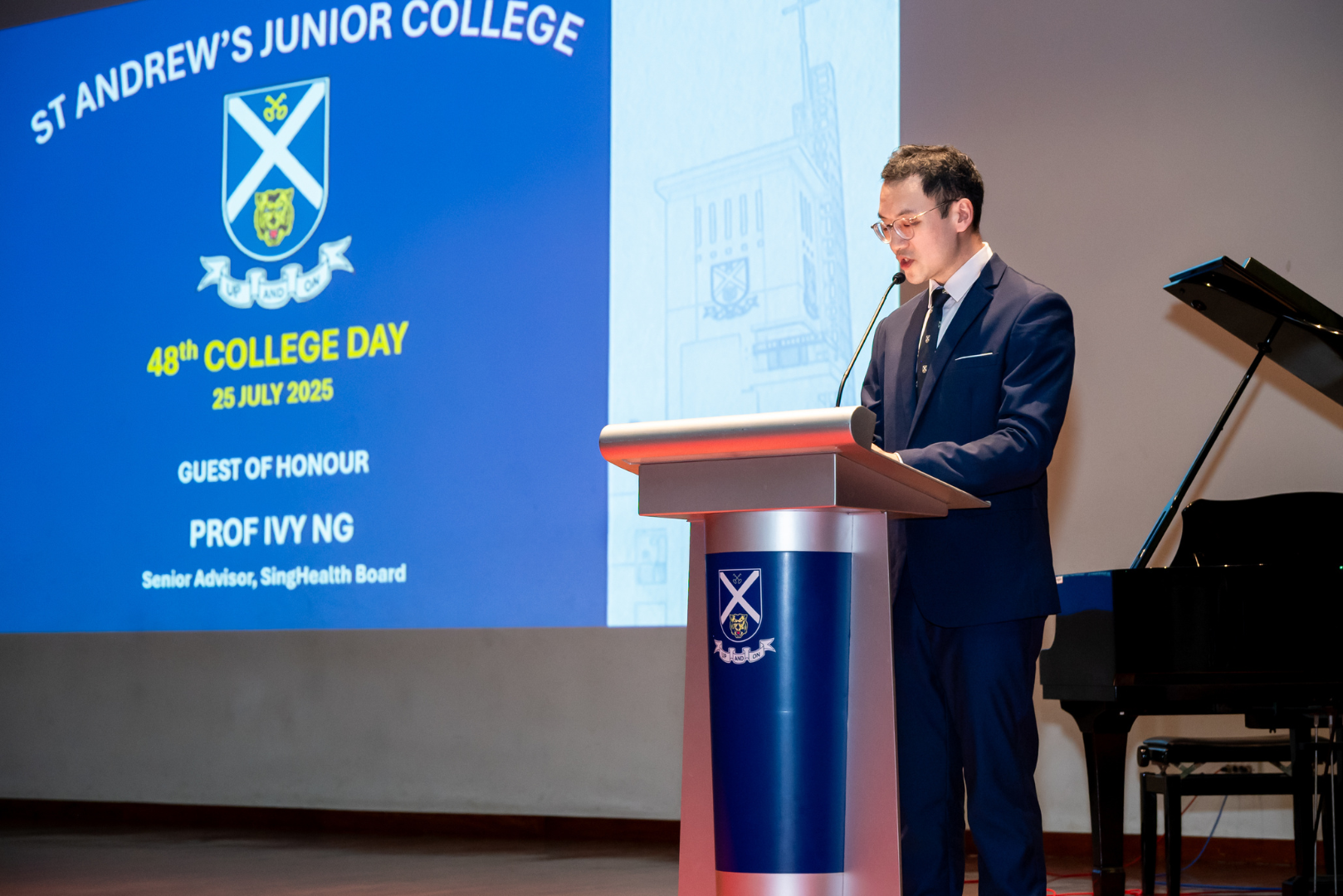 Mr Wayne Ko speaks at a podium bearing the St Andrew's Junior College crest. Behind him is a large projection screen displaying the event details: St Andrew's Junior College, 48th College Day, 25 July 2025, with the Guest of Honour listed as Prof Ivy Ng, Senior Advisor, SingHealth Board. A grand piano is visible to the right of the stage.