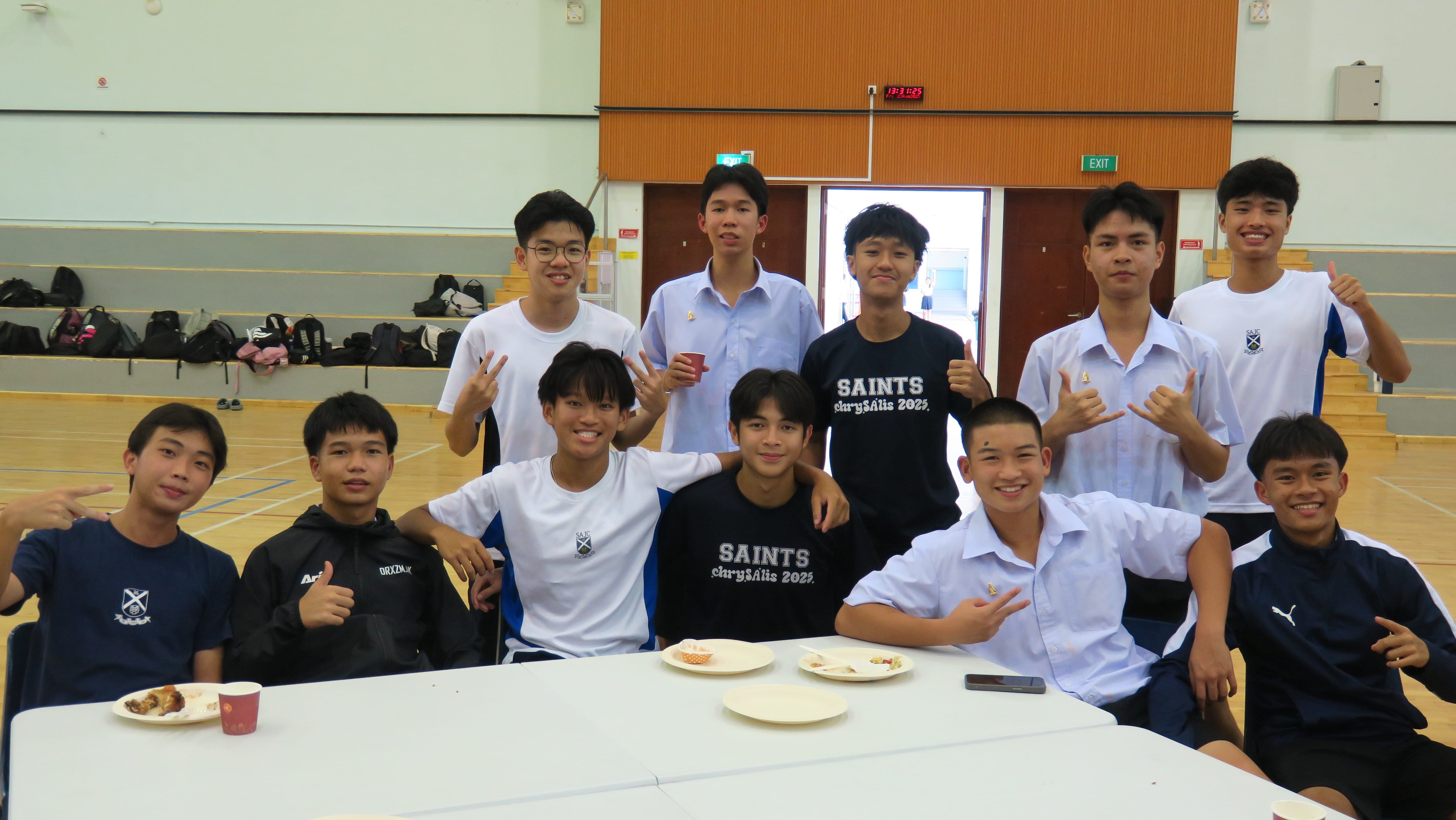 A group of male students from both St Andrew's Junior College and Triam Udom Suksa School pose cheerfully around a table in a school hall, giving thumbs up and peace signs after a meal.