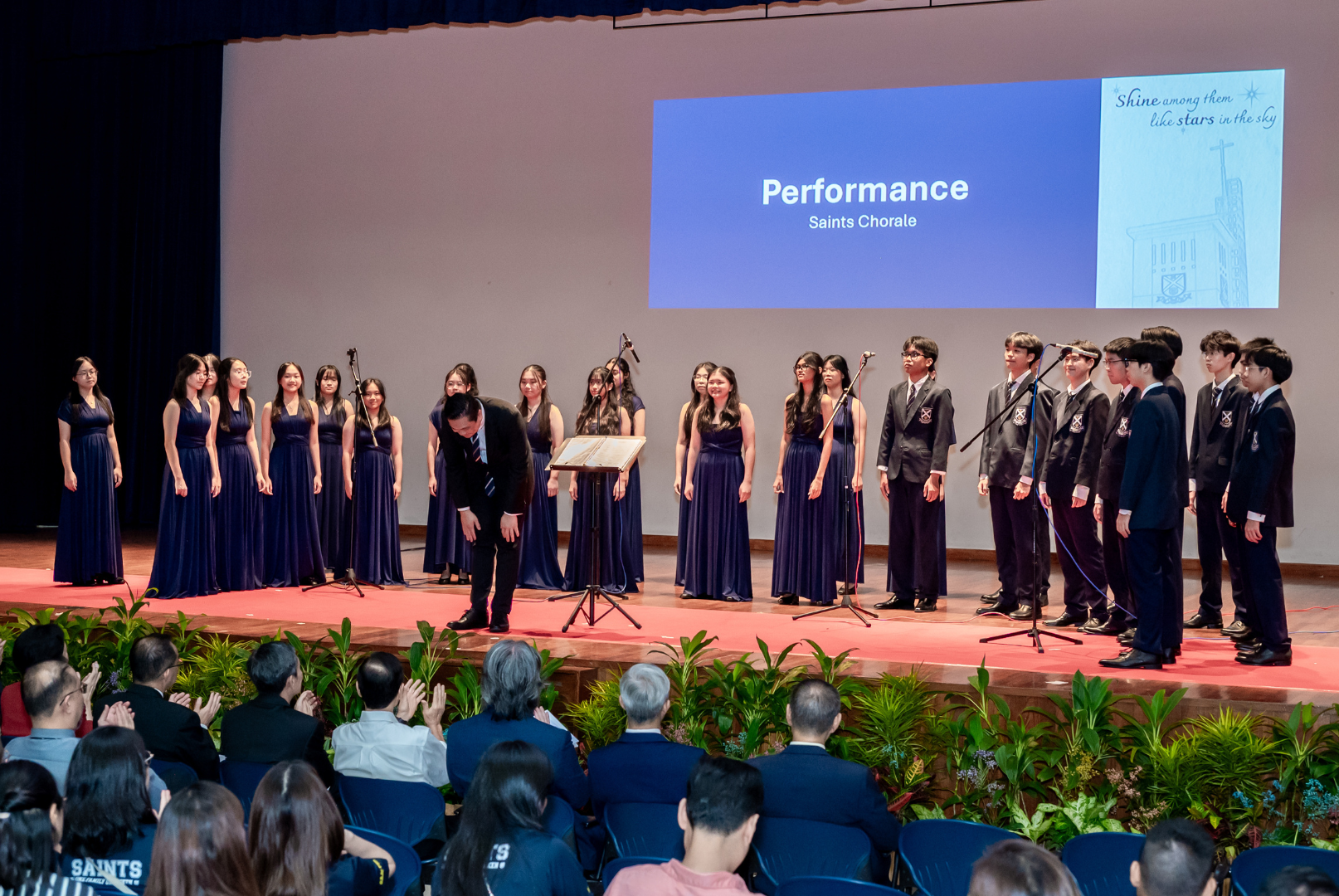 The Saints Chorale performs on stage at St Andrew's Junior College, with female members in navy gowns and male members in school blazers, as their conductor takes a bow before the audience. A slide reading 'Performance - Saints Chorale' is projected on screen behind them.