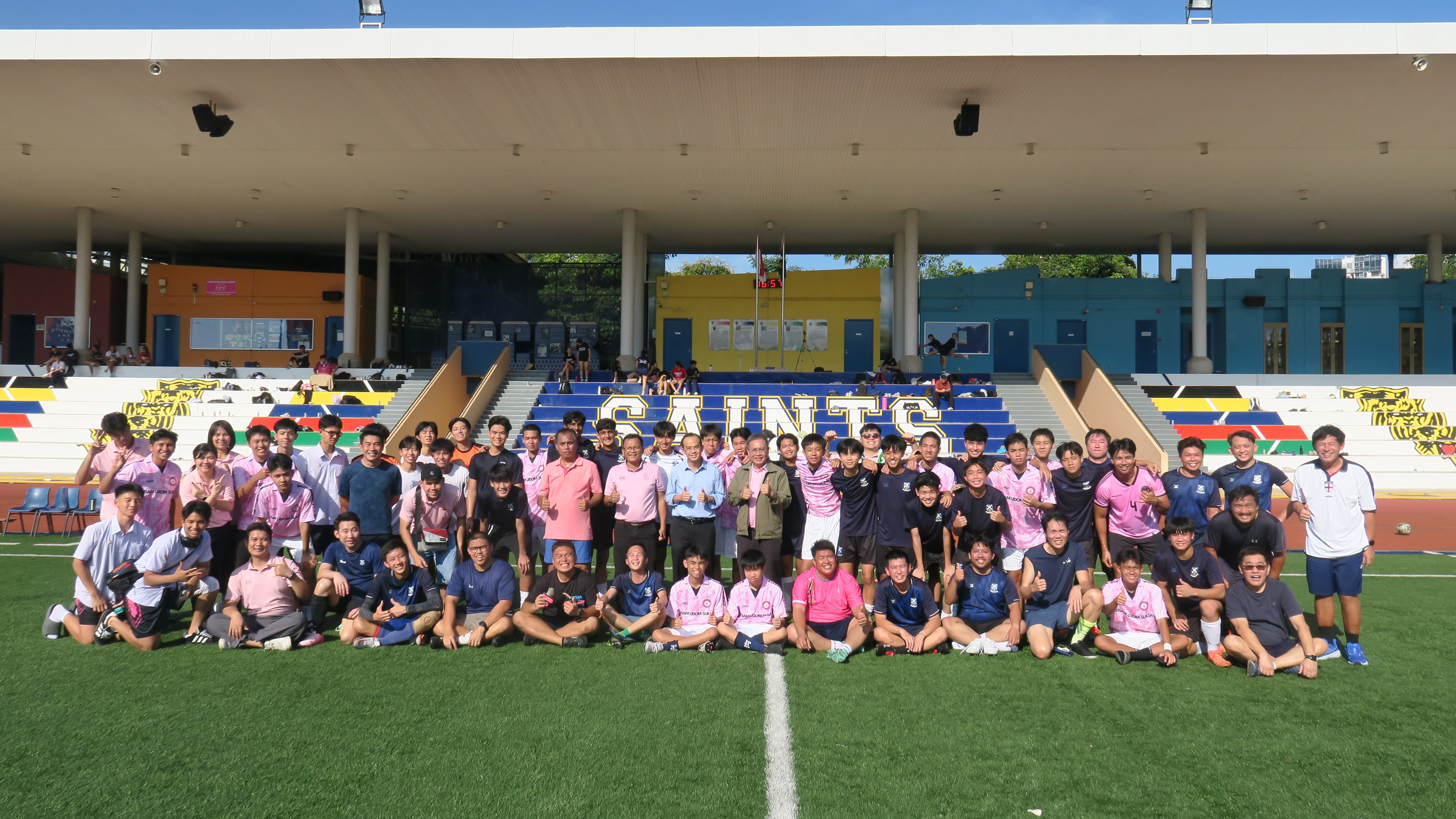 Large group in sports jerseys posing on a soccer field, with colorful bleachers and a covered stand behind.