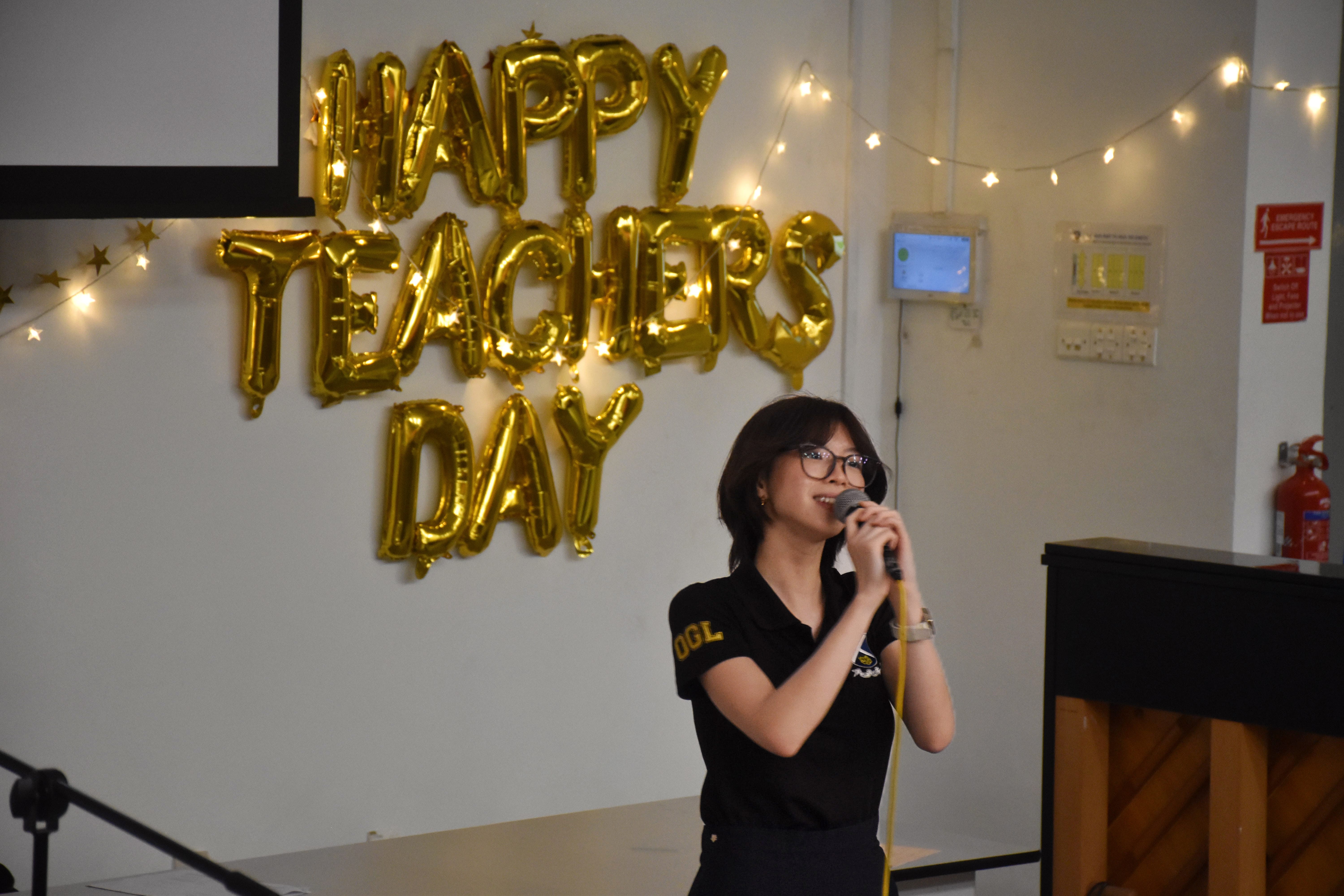 A smiling young woman wearing glasses and a black polo shirt with "OGL" written on the sleeve speaks into a microphone. Behind her on a white wall, gold metallic balloons spell out "HAPPY TEACHERS DAY," framed by warm fairy lights.