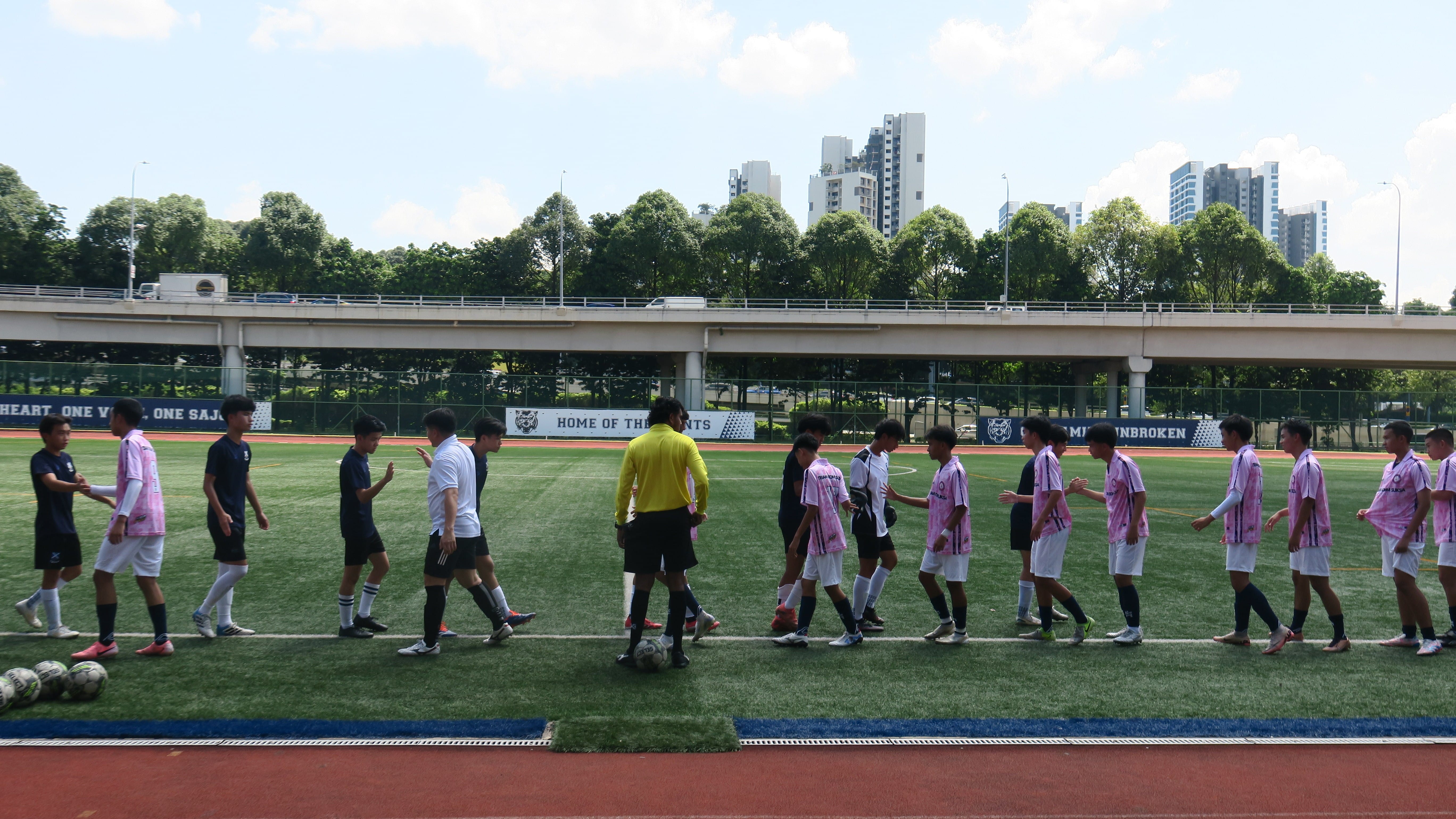 Two football teams shake hands on a green pitch, with a referee in yellow standing between them and a school stadium banner reading "Home of the Saints" in the background.