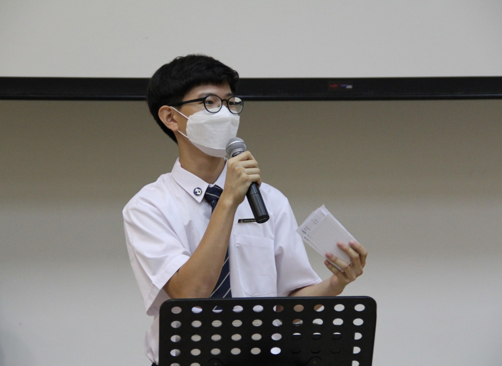 A student in school uniform wearing a face mask and glasses speaks into a microphone while holding notes, standing at a music stand.