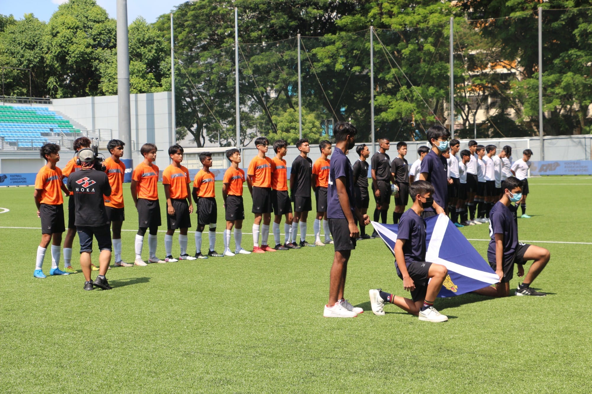 SAJC Boys' Football team lined up on the pitch alongside their opponents before a match, with a student kneeling in the foreground holding the SAJC flag
