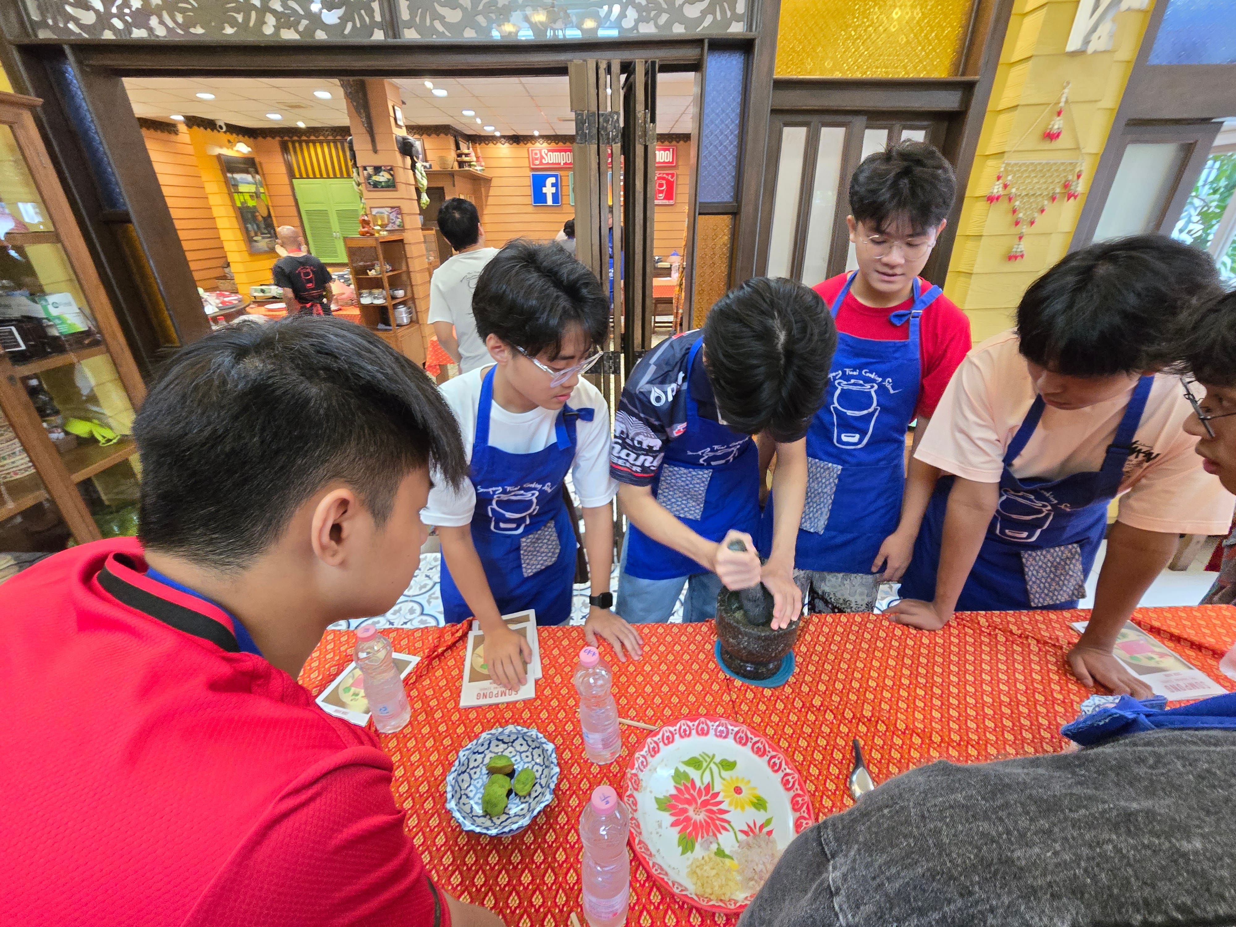 Students wearing blue aprons gather around a table at a Thai cooking class, with one student using a mortar and pestle to grind ingredients. Plates of food and water bottles are visible on the orange-patterned tablecloth.