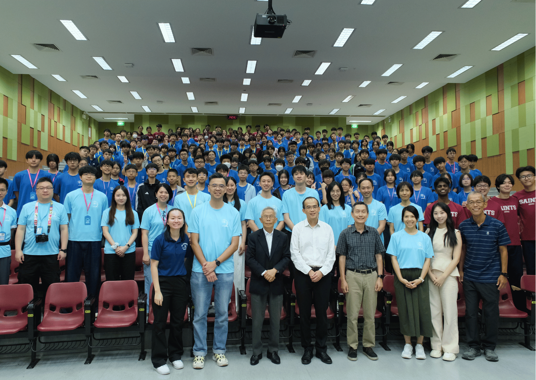 A large group photo taken in a lecture theatre, Tsung Tsin Christian Academy students in blue event T-shirts alongside staff and distinguished guests in formal attire.