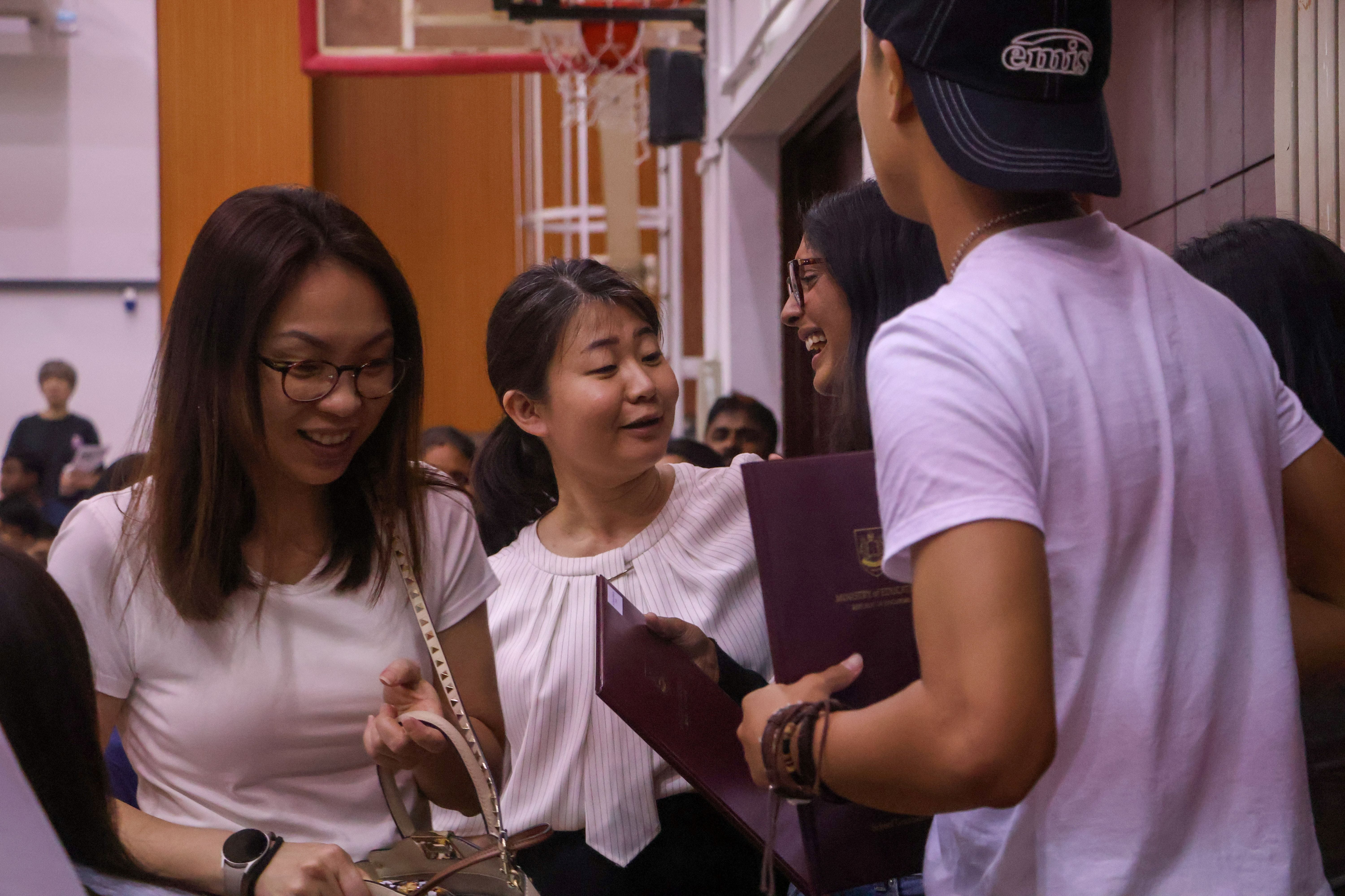 Three smiling young people talking indoors, one holding a dark red official certificate folder.