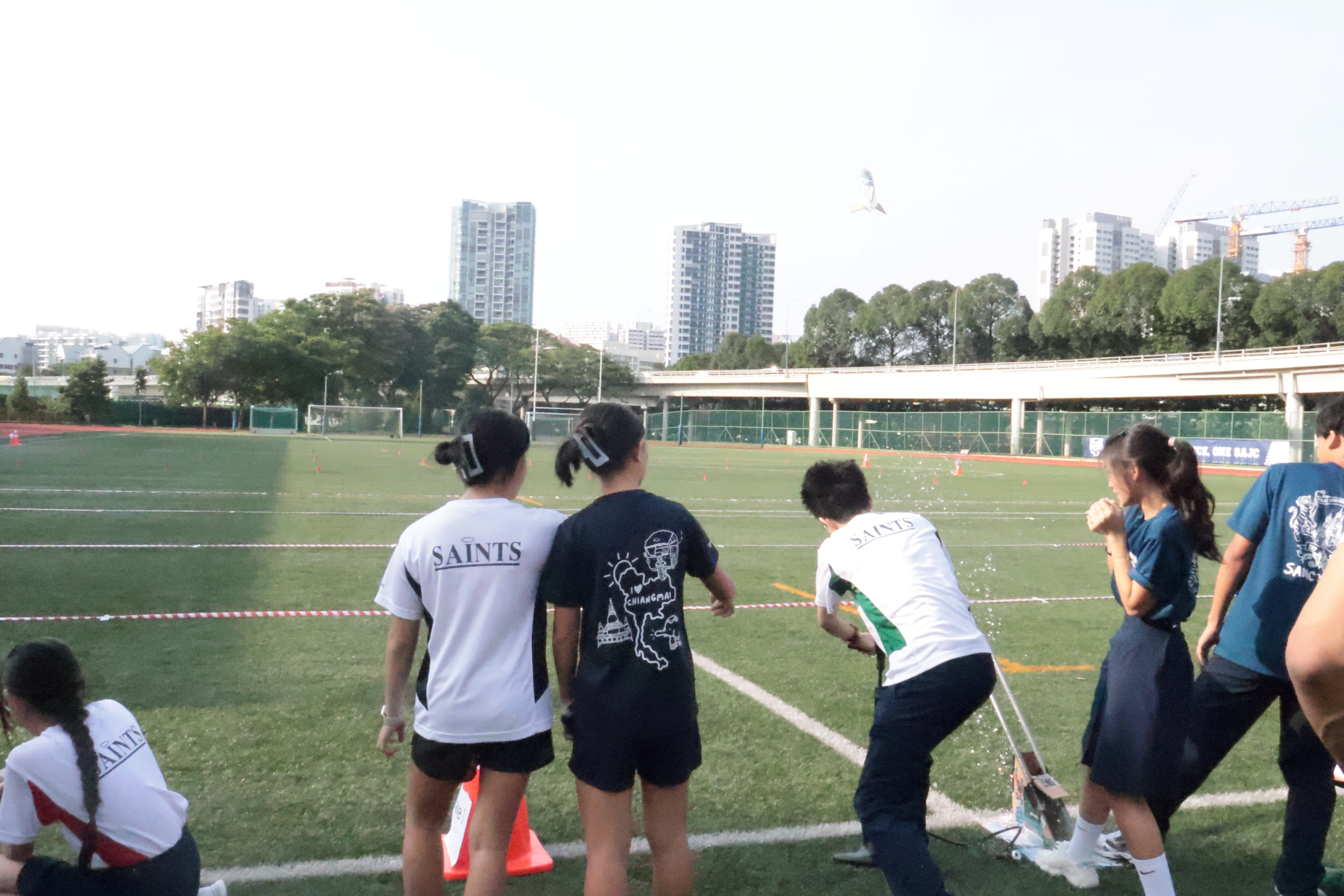 Students in Saints shirts stand on a green sports field with city buildings visible in the background.