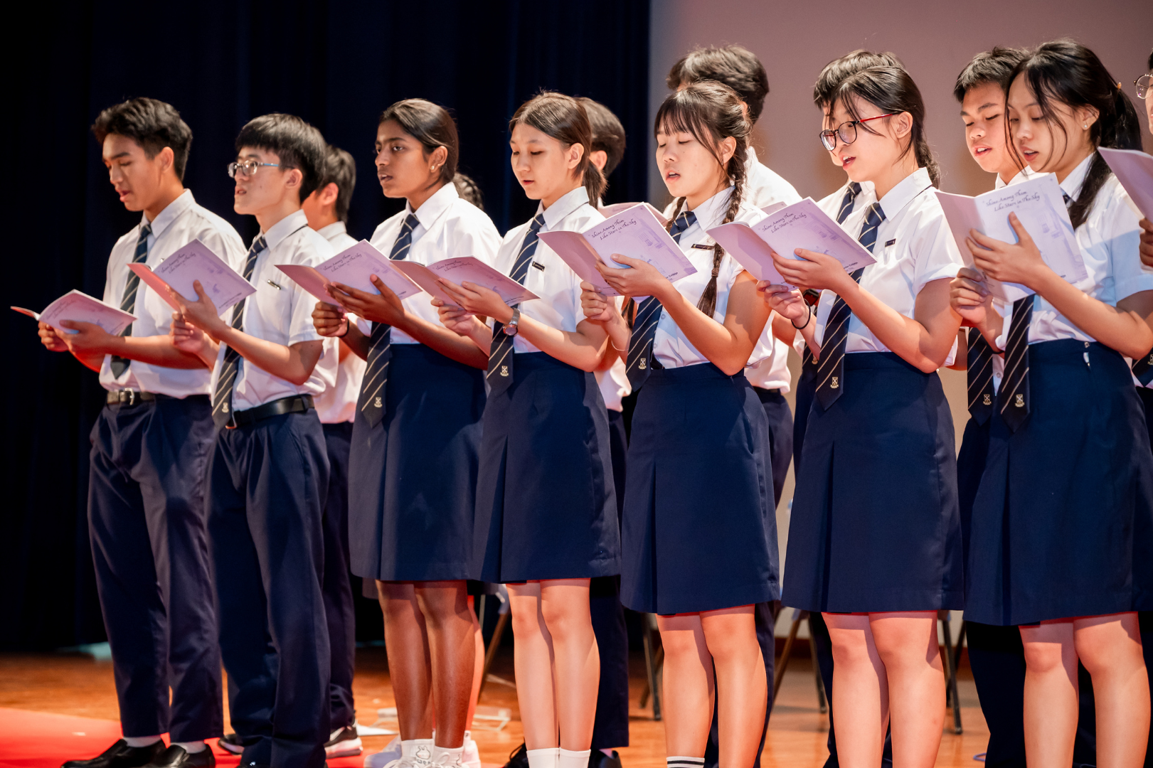 A group of students in school uniforms stand in a row on stage, each holding and reading from pink booklets, appearing to perform as a choir or recitation group.
