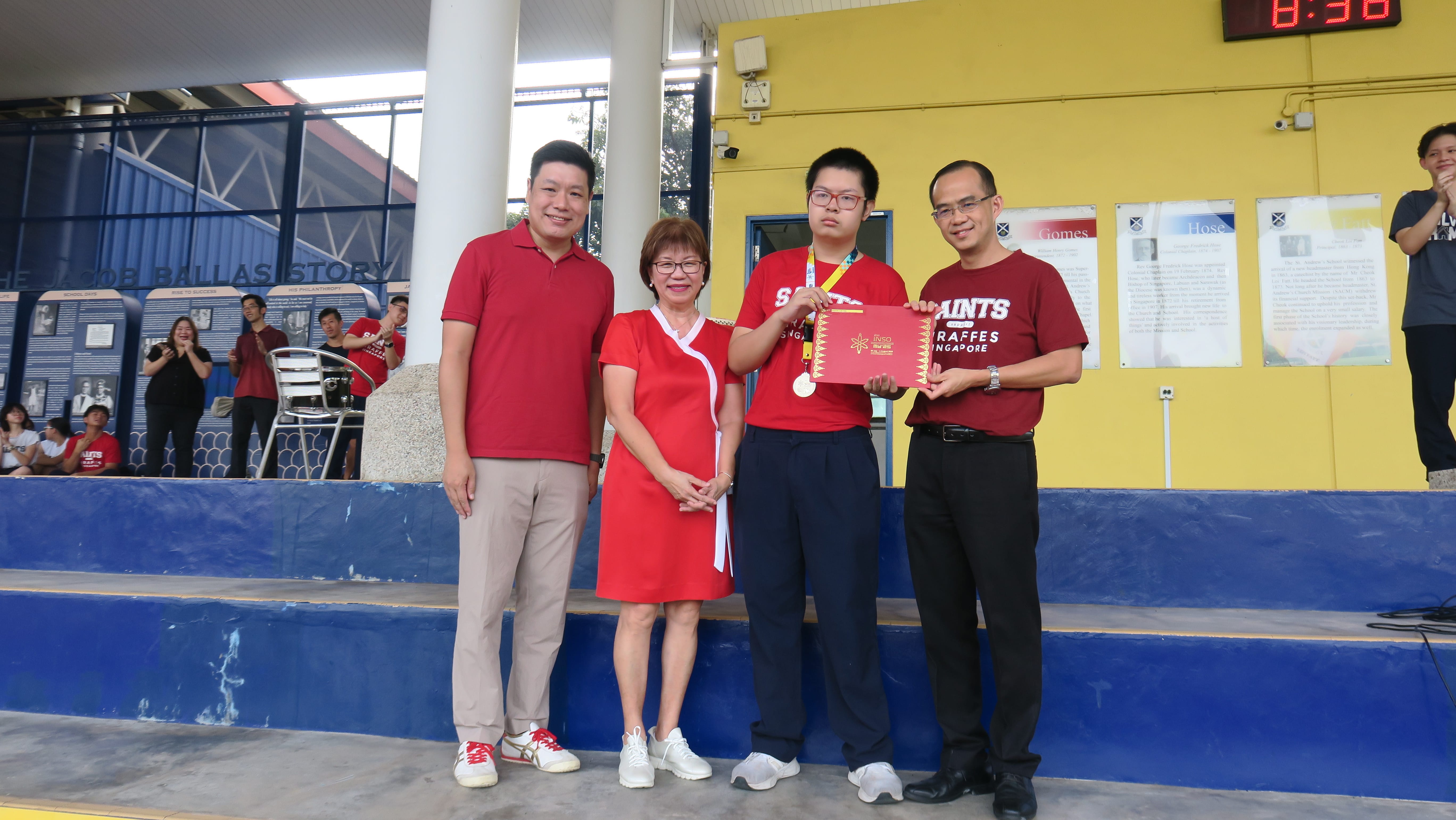 Four people pose outdoors; a student wearing a medal holds a red certificate alongside two adults in red shirts and a woman.