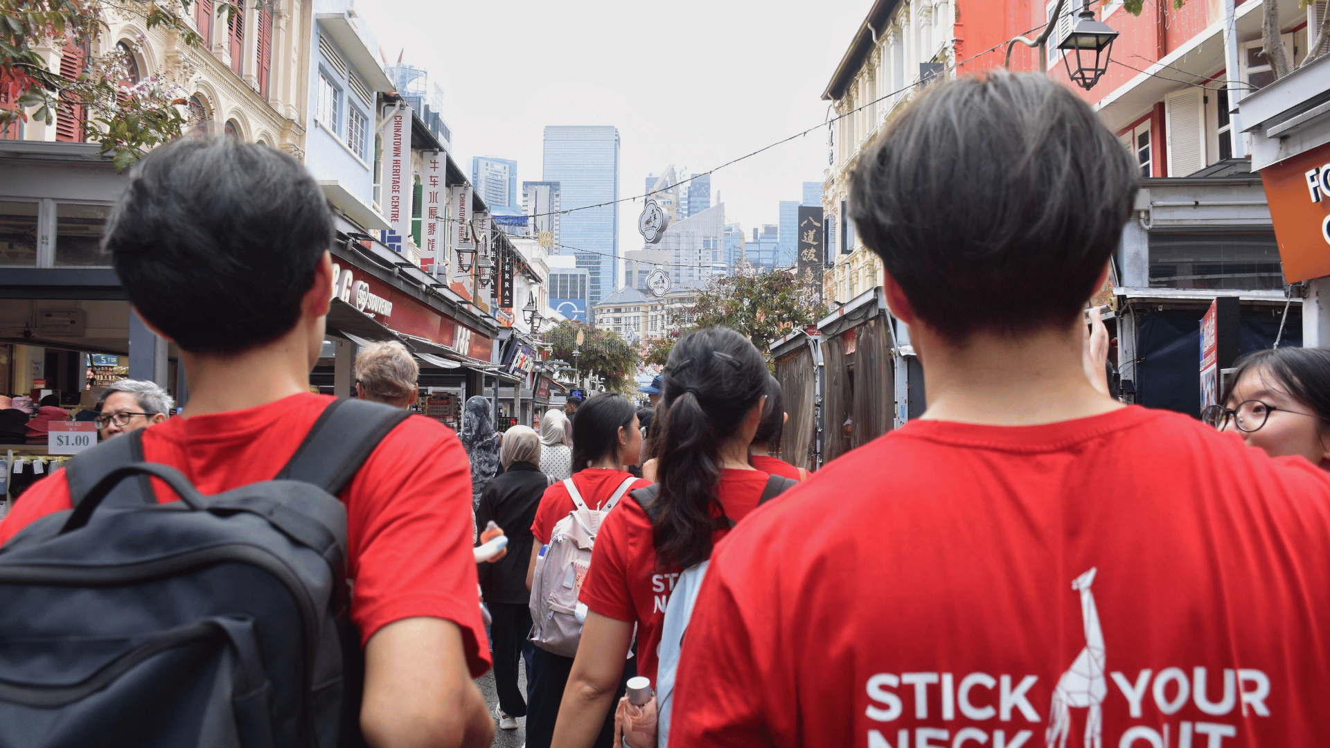 A rear-view photo of individuals wearing bright red t-shirts walking along a pedestrianized street lined with traditional shophouses. The back of the shirt in the right foreground features a white giraffe silhouette and text that reads "STICK YOUR NECK OUT." Down the center of the street, shop signboards (including one for Chinatown Heritage Centre) line the heritage buildings, with modern city skyscrapers visible in the background under an overcast sky.