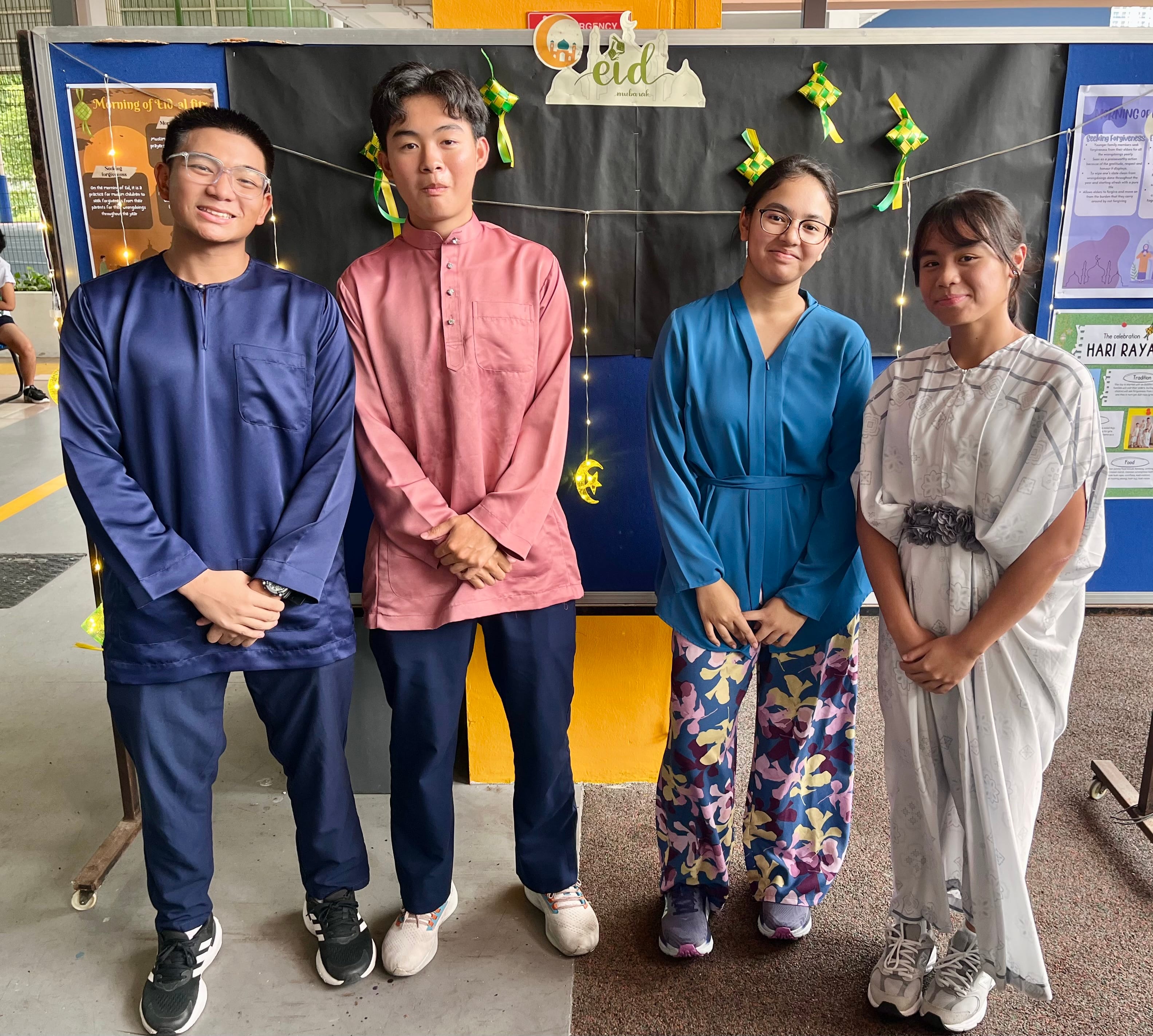 4 students in Malay ethnic attire posing in front of a noticeboard with announcements regarding the celebration of Hari Raya