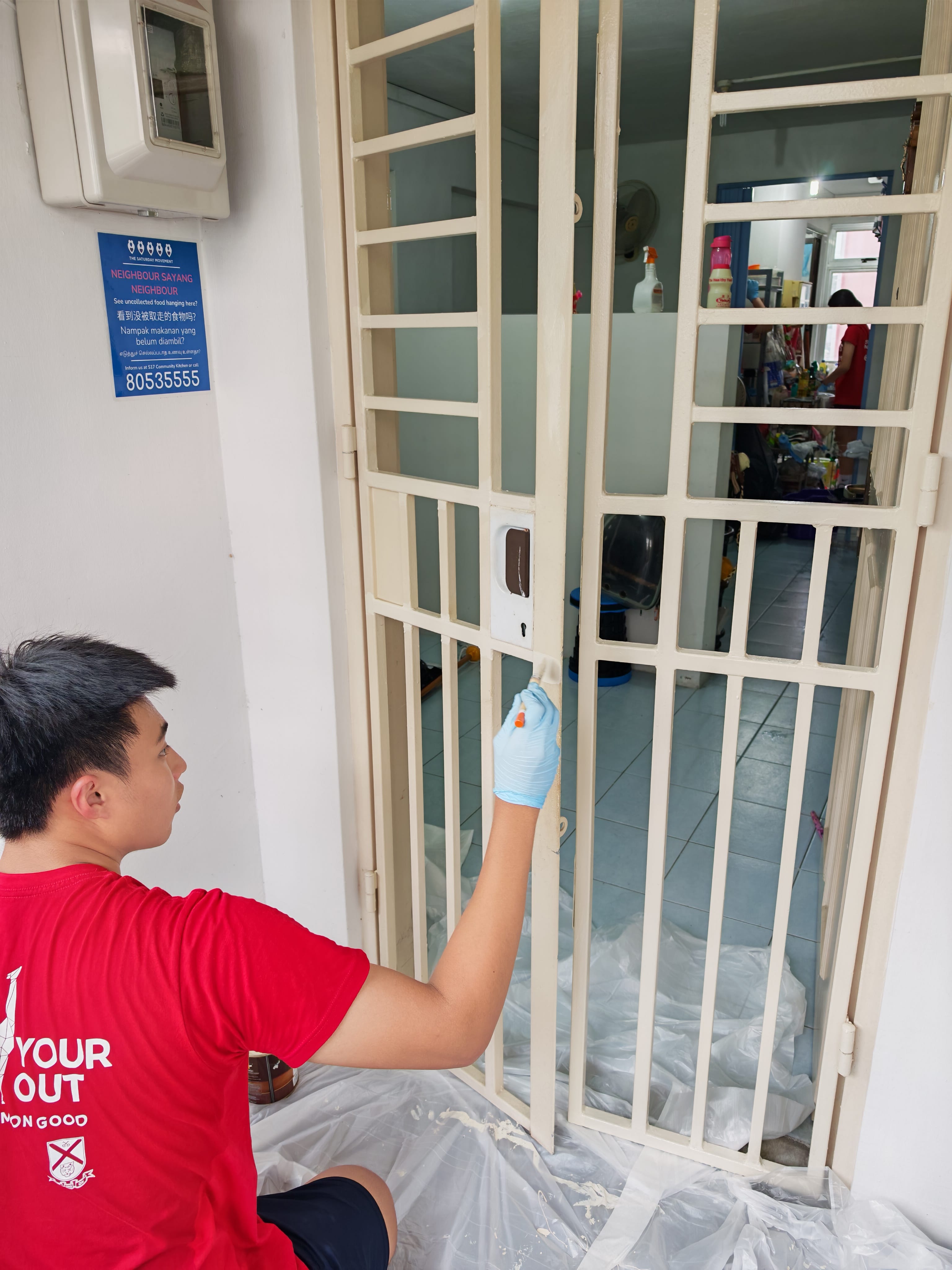 A young male volunteer wearing a red t-shirt and blue disposable gloves crouches outside a residential unit, carefully applying paint to a metal gate with a small brush. Clear plastic sheeting covers the floor below to catch drips, and a blue neighborhood poster is displayed on the wall next to an electrical meter box.