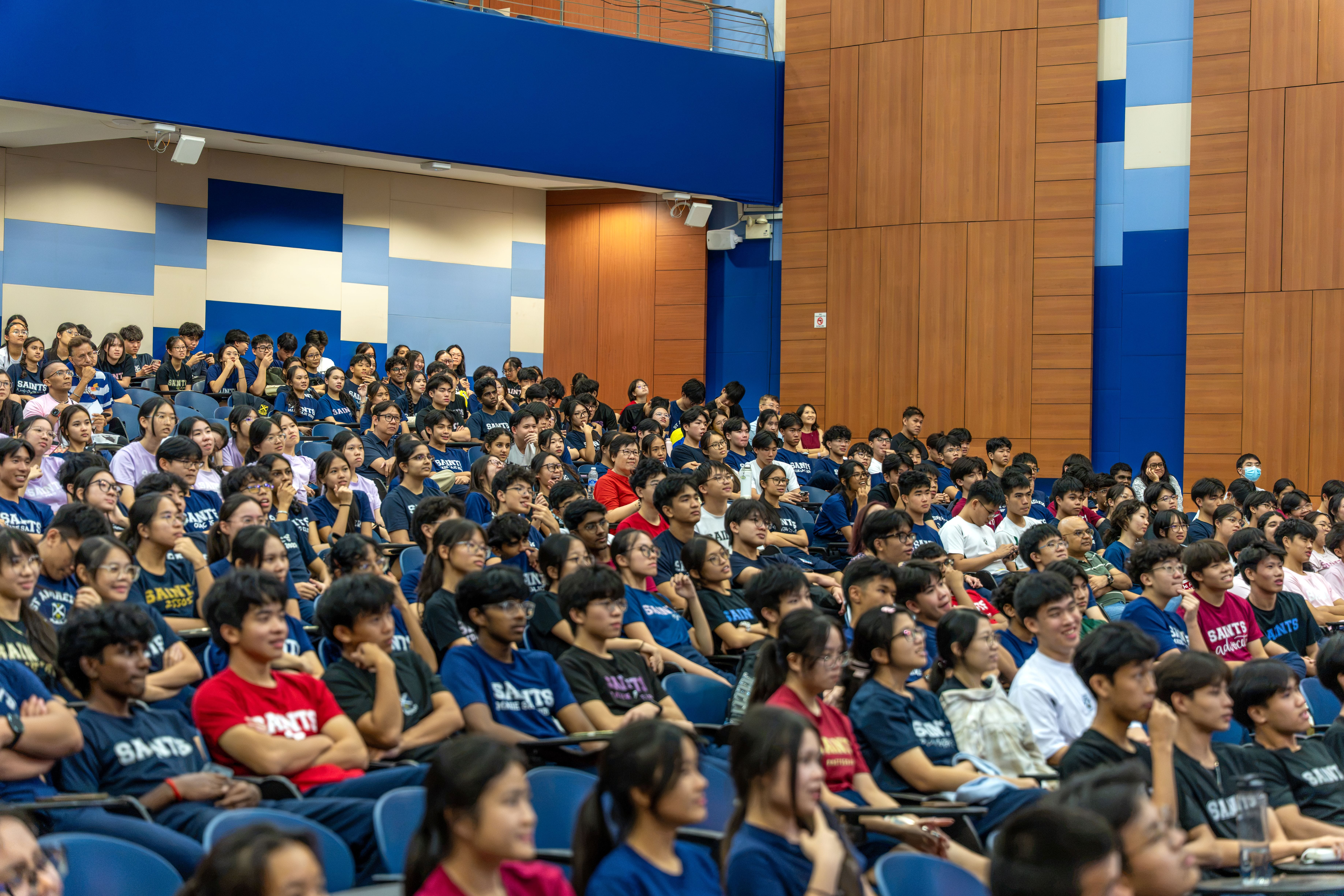 A wide shot of a spacious lecture hall filled with a crowd of seated students. Most attendees are wearing blue or red t-shirts with "SAINTS" printed on them. The audience faces forward with attentive expressions, surrounded by wood-paneled and blue accent walls in a multi-tiered seating arrangement.