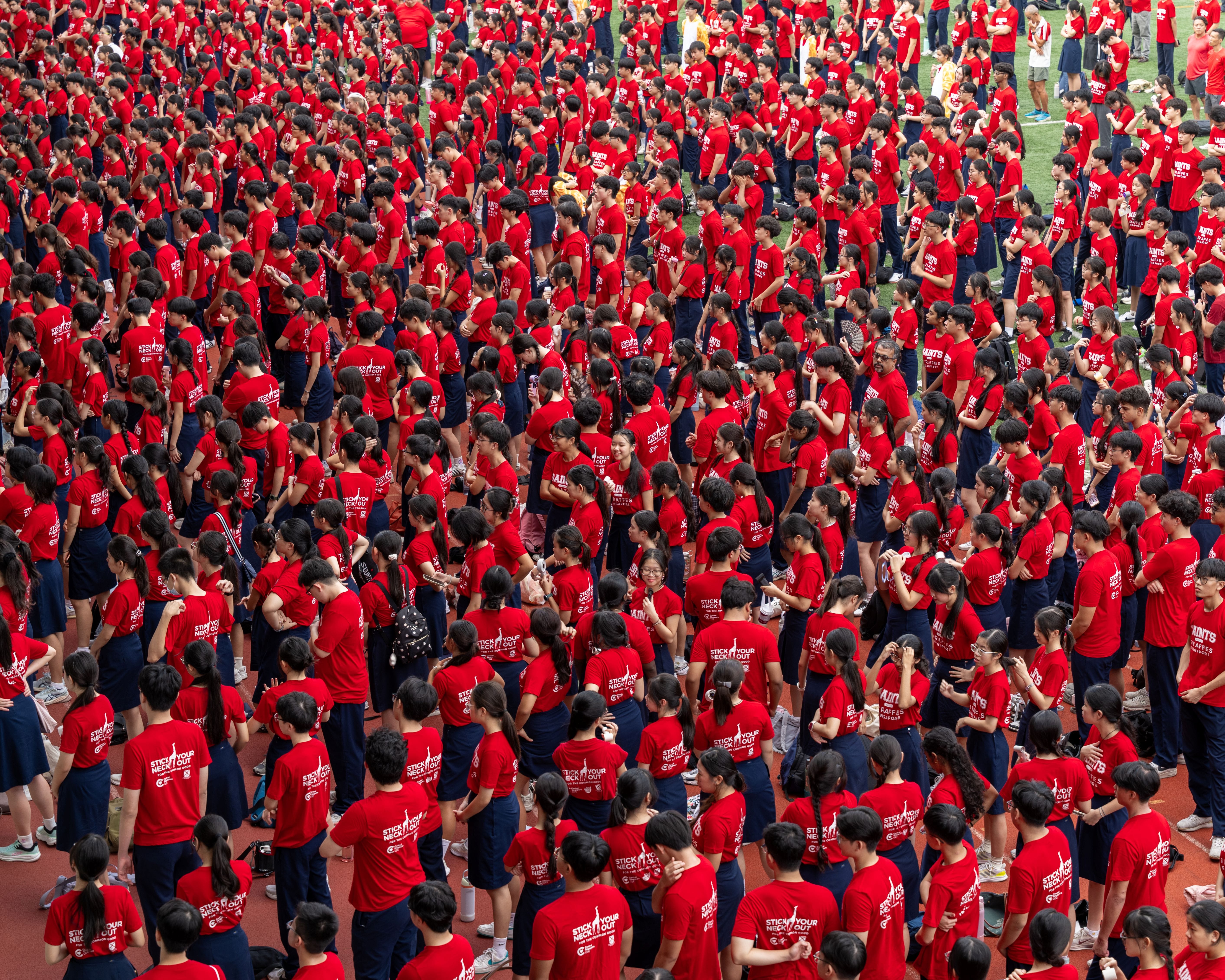 An elevated, high-angle photograph showing a massive crowd of students gathered tightly together on an outdoor running track surface. Almost everyone is dressed in matching red short-sleeved t-shirts paired with dark blue skirts, shorts, or trousers. The backs of several t-shirts feature a white giraffe silhouette graphic with the slogan "STICK YOUR NECK OUT." The sea of red-clad individuals extends densely from the foreground deep into the background.