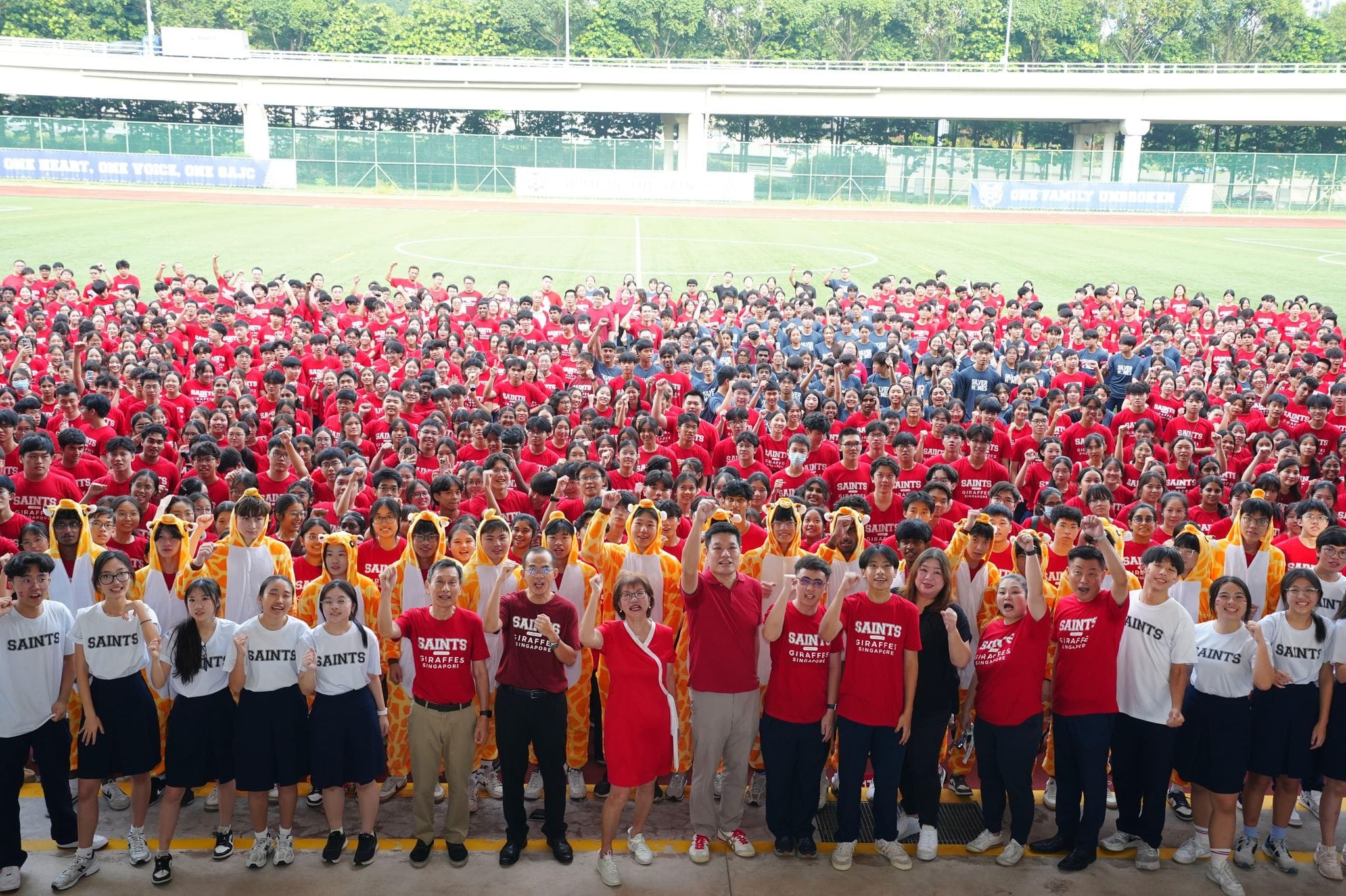 Large group of students in red and white Saints shirts pose cheering at a sports field, some wearing giraffe costumes.