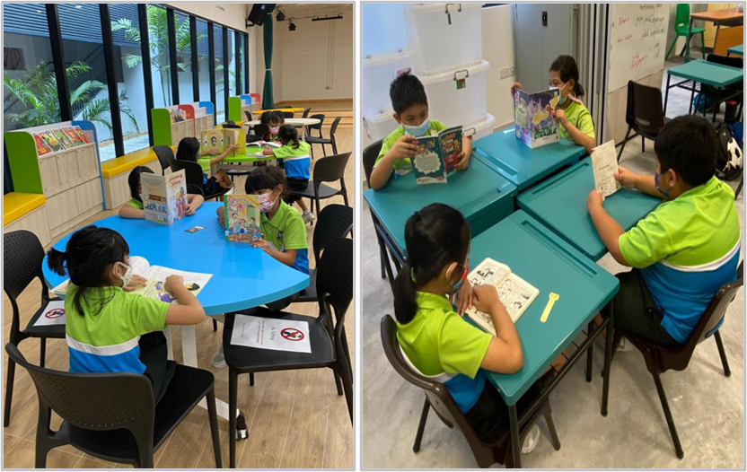 Children in green uniforms and masks sit at blue tables reading books in a bright classroom library.