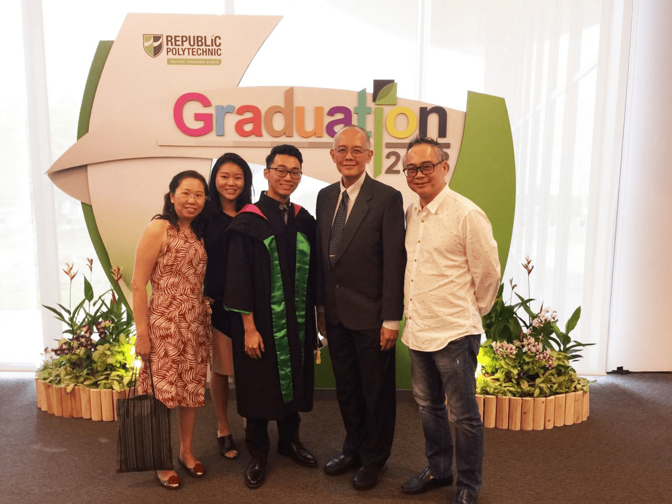 Five people pose in front of a Republic Polytechnic Graduation sign, one wearing a black academic gown with green stole.