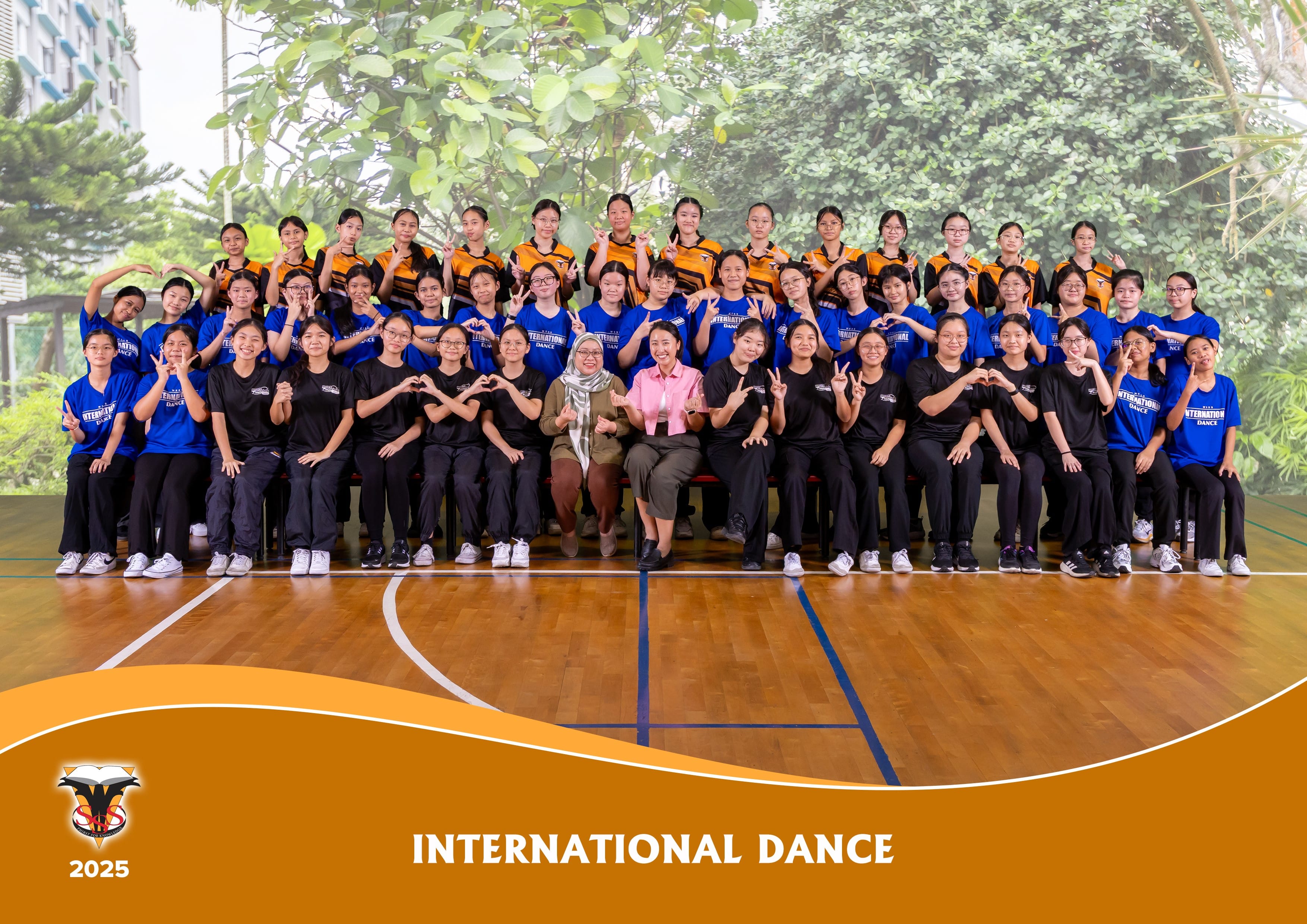 Large group of students in blue and orange shirts posing on a gym floor for International Dance 2025 team photo.