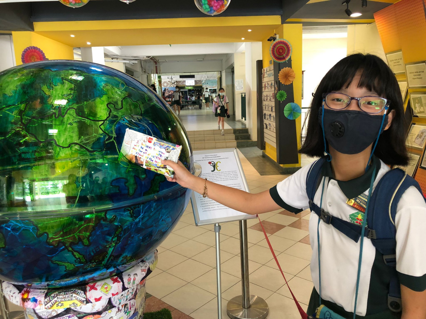 A masked girl with glasses holds a paper against a large decorative globe display in a colorful school hallway.