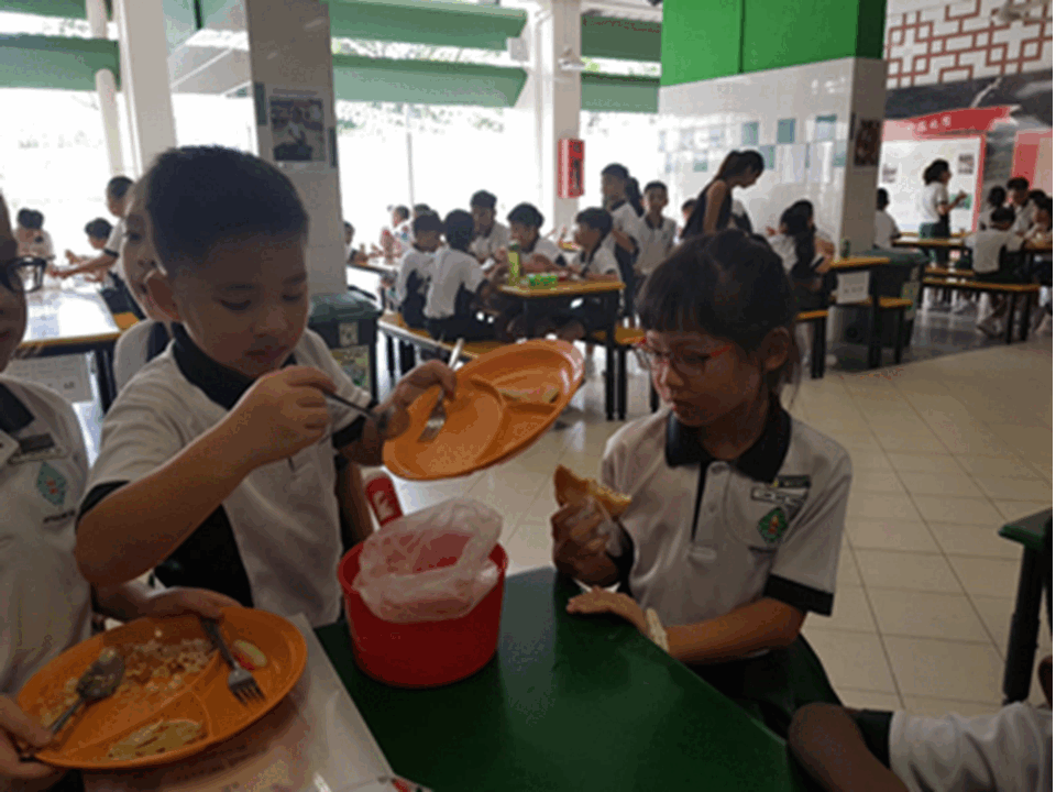 Two school children in uniforms eating lunch at a green table in a busy school canteen.
