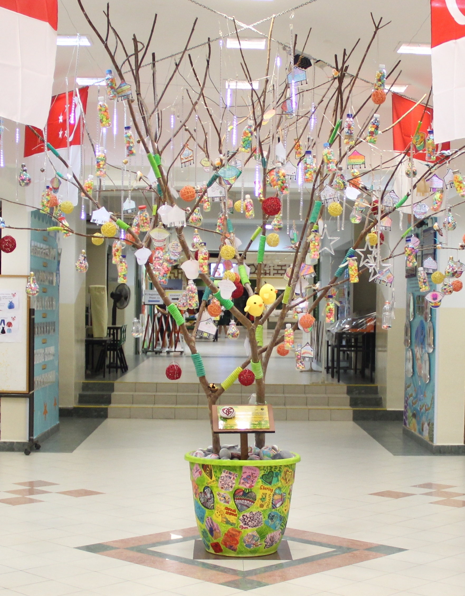 Bare tree in a colorful pot decorated with hanging ornaments, bottles filled with candies, and paper cutouts indoors.