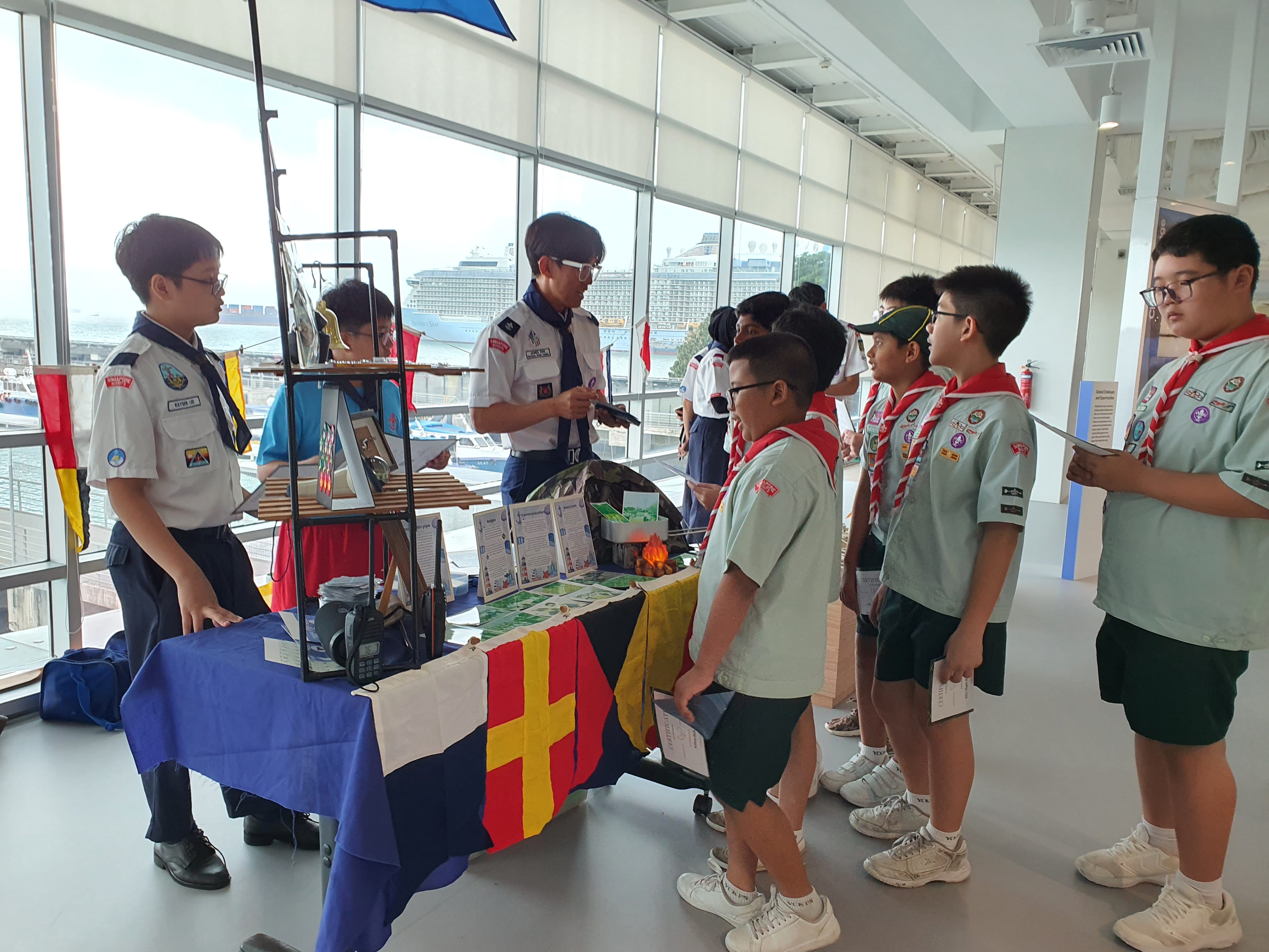 Boy scouts in uniforms with red scarves stand at a display table with flags and materials near large windows overlooking a harbor.