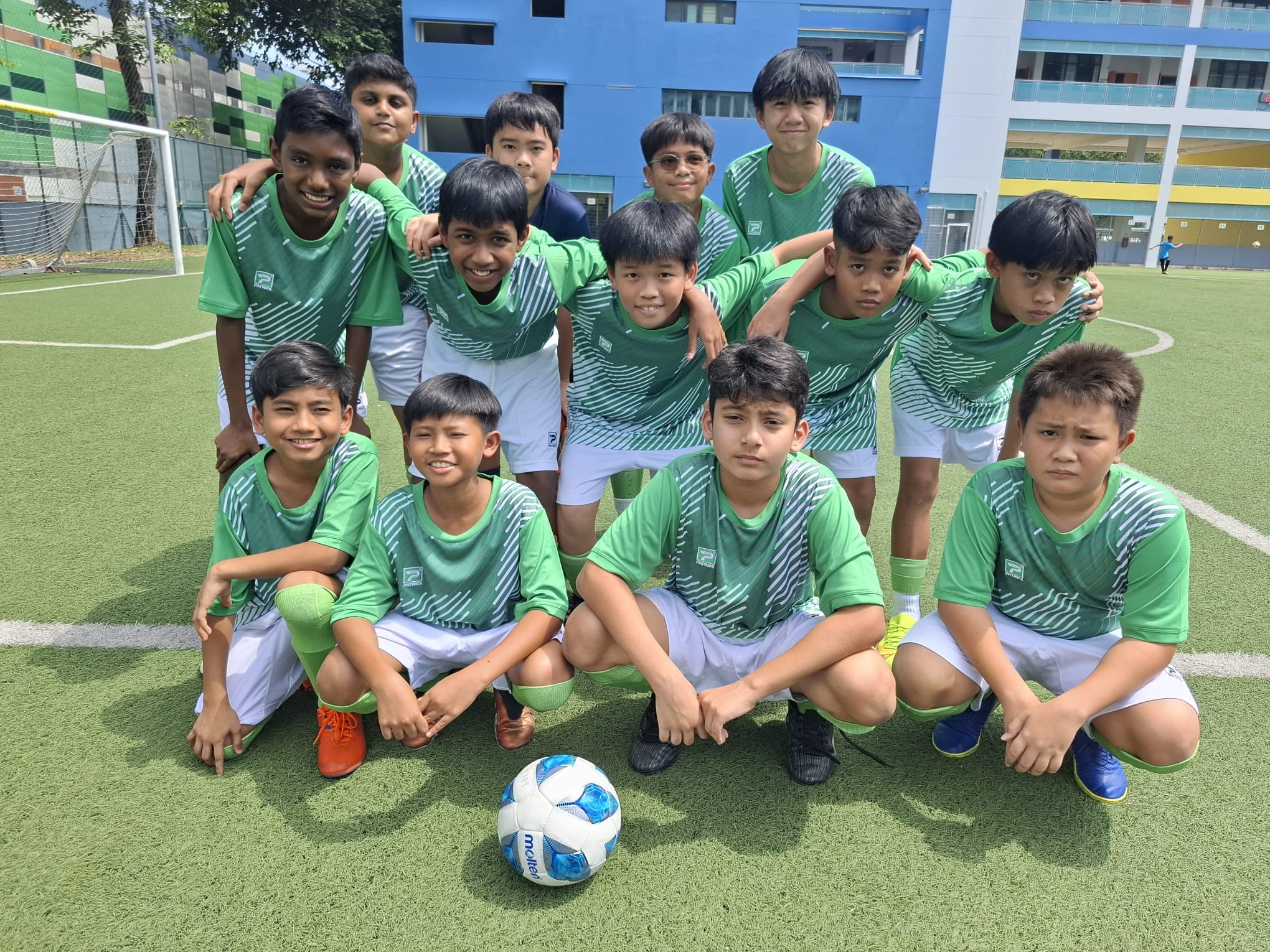 A youth soccer team of about 12 boys in green uniforms posing on a turf field with a soccer ball in front.