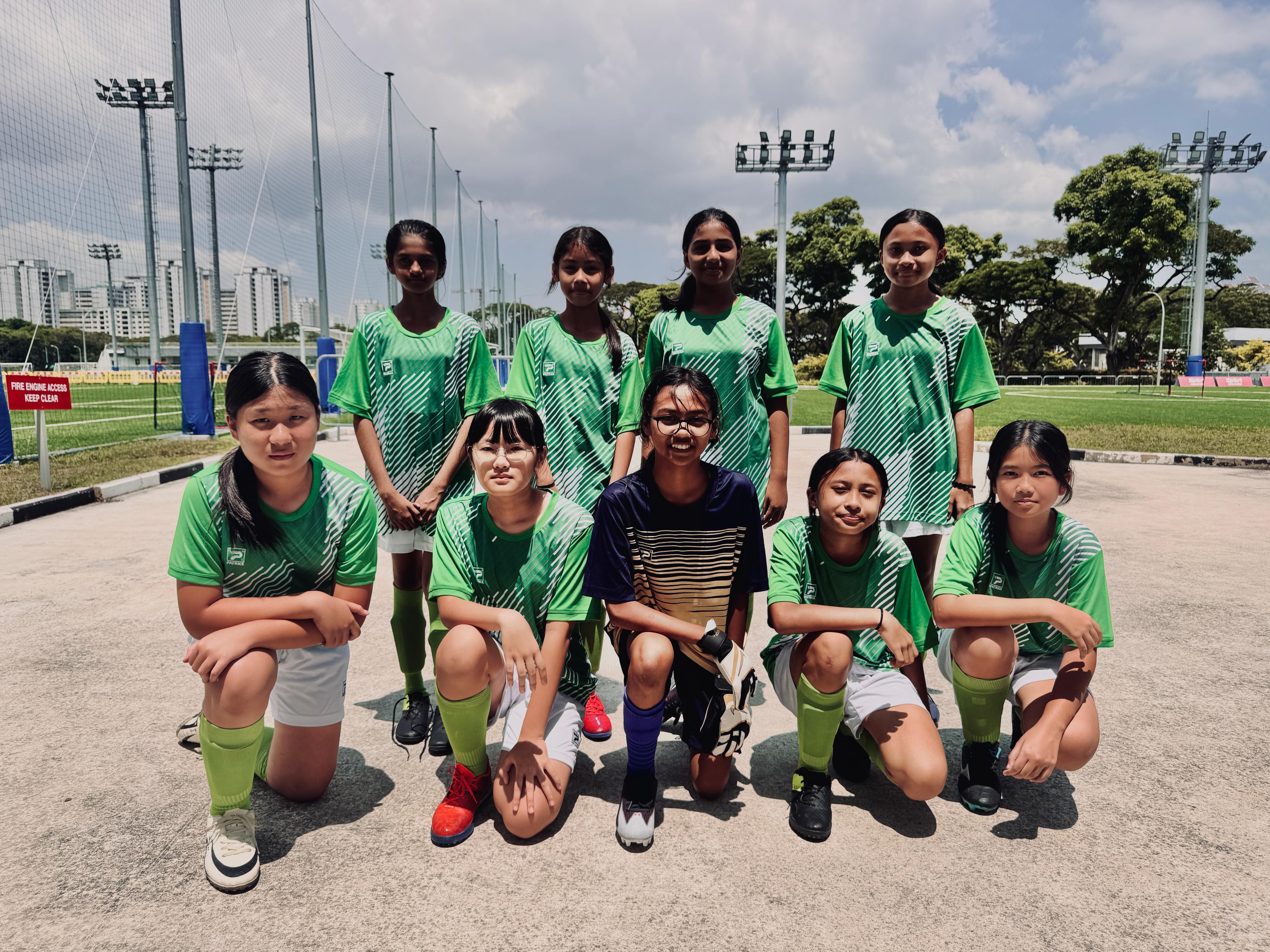 A girls soccer team in green jerseys poses in two rows at an outdoor sports field with floodlight poles.