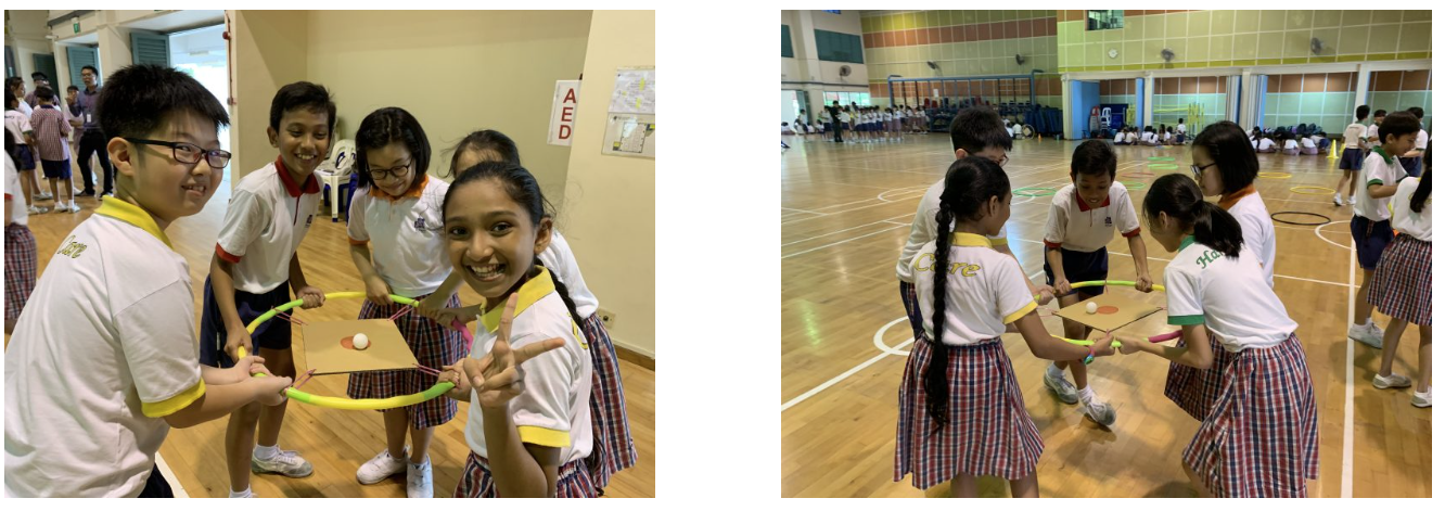 Students in school uniforms balance a ball on a cardboard sheet using hula hoops in a gymnasium activity.