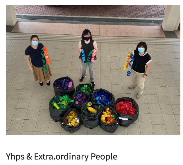 Three masked people stand outdoors holding colorful ribbons, with several black bags full of colorful plastic items on the ground.