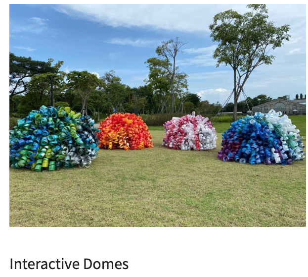 Four colorful dome sculptures made from recycled plastic bottles sit on a grassy field under a blue sky.