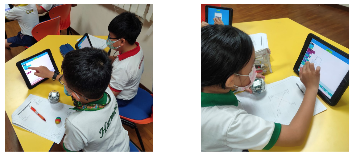 Students wearing masks use tablets at yellow desks with worksheets and small robotic balls nearby.