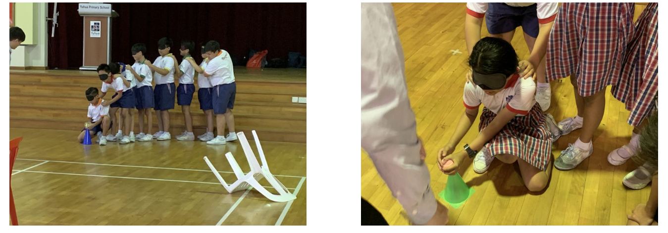 Children in school uniforms play a blindfolded team activity on a wooden gym floor with small cones.