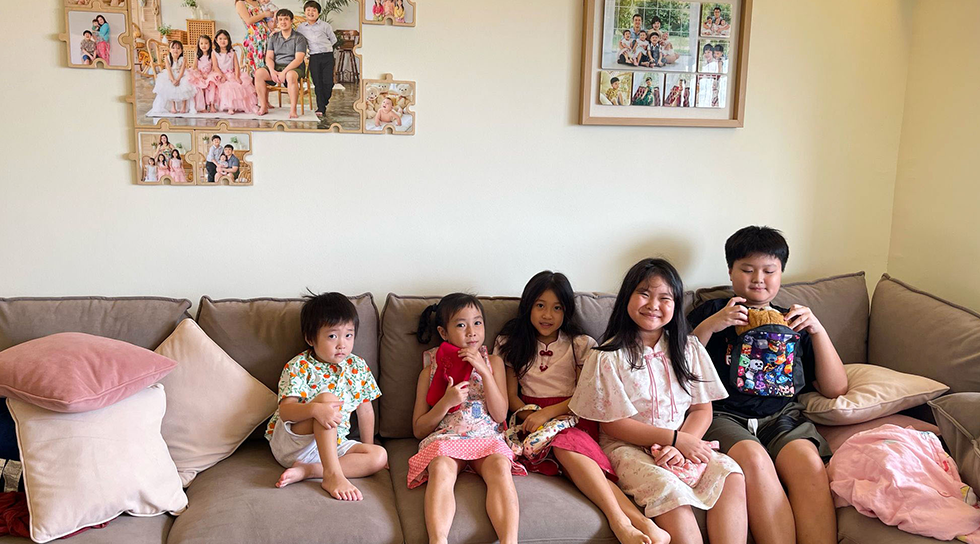 Five children sitting together on a sofa beneath framed family photos at home