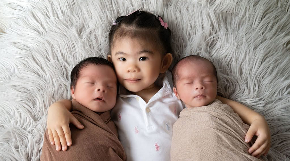 Young girl lying beside newborn twins wrapped in blankets on a soft grey rug