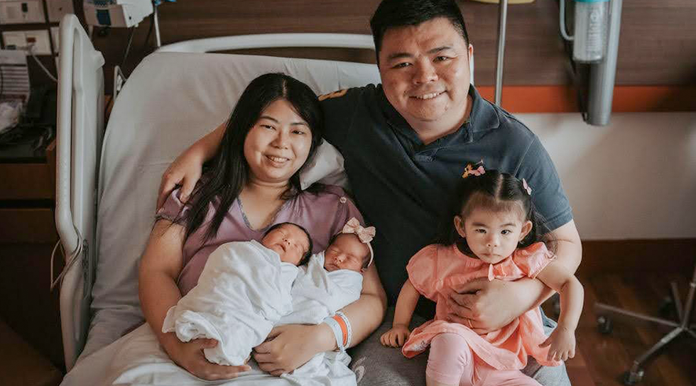 Parents with newborn twins and a young child posing together in a hospital room