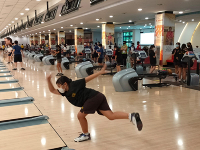 A masked bowler releases a ball on a busy indoor bowling alley with many lanes and players in the background.