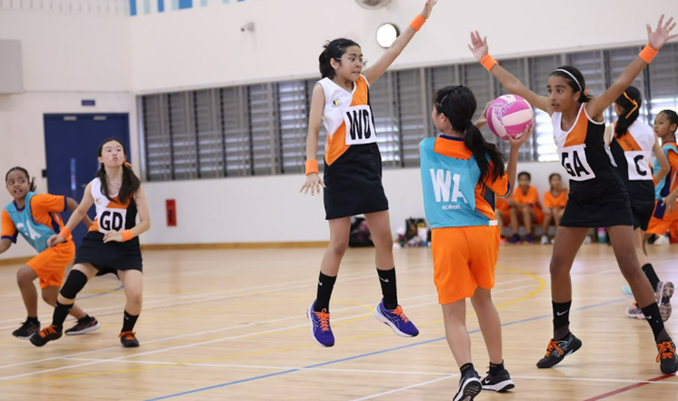 Girls playing netball indoors, one player jumping with arms raised while another holds a pink and white ball.
