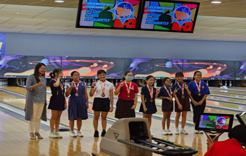Group of young girls wearing medals posing and smiling at a bowling alley with colorful screens above.